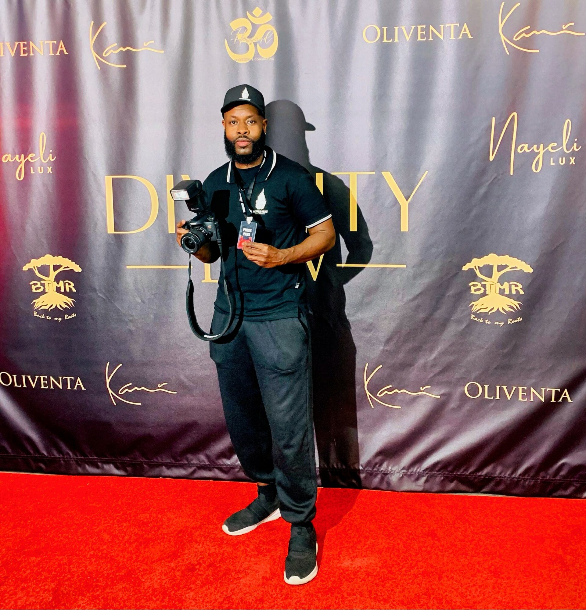 Man standing on red carpet holding camera, black backdrop with gold and white logos behind him.