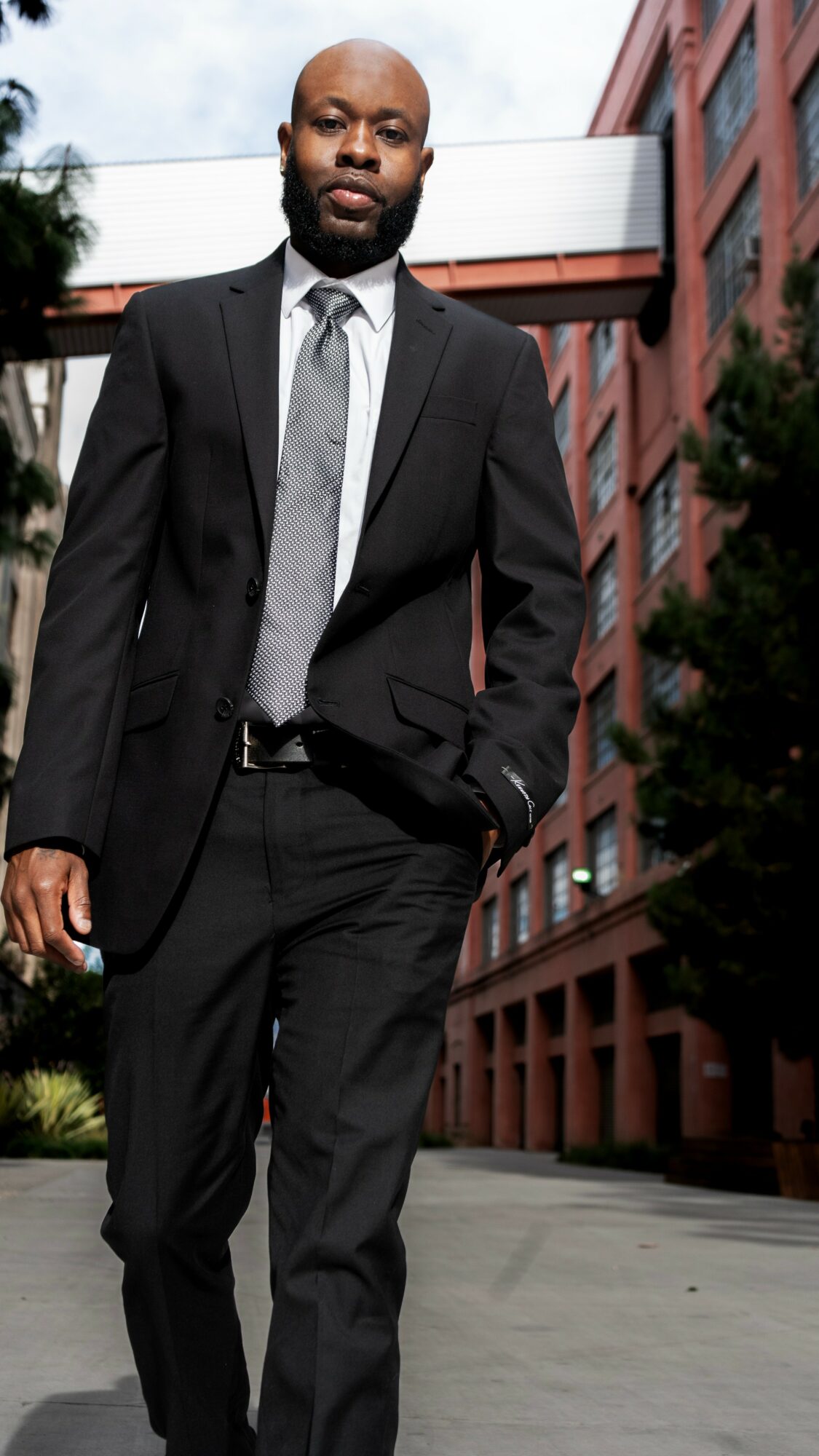 Man in suit walking outdoors near red brick building, trees, and sidewalk.