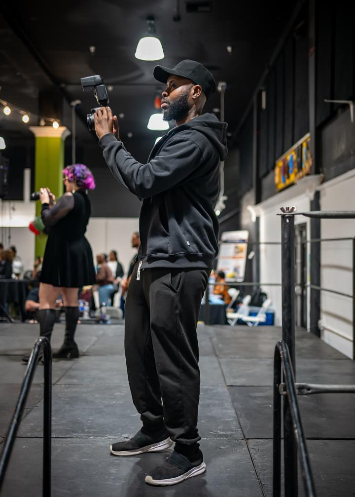 Man in black hoodie and pants holding a camera in a busy indoor space with people and colorful decorations.