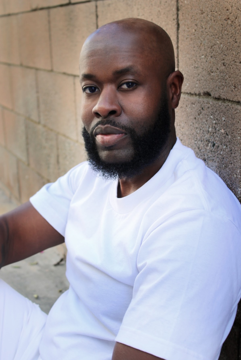 Man with a beard and shaved head wearing a white shirt, sitting against a brick wall.