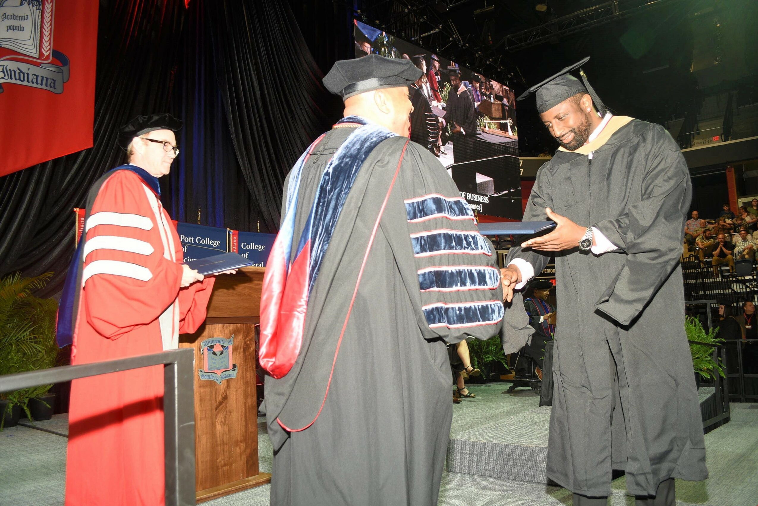 Person in graduation gown and cap receiving diploma from another person in academic regalia on stage.