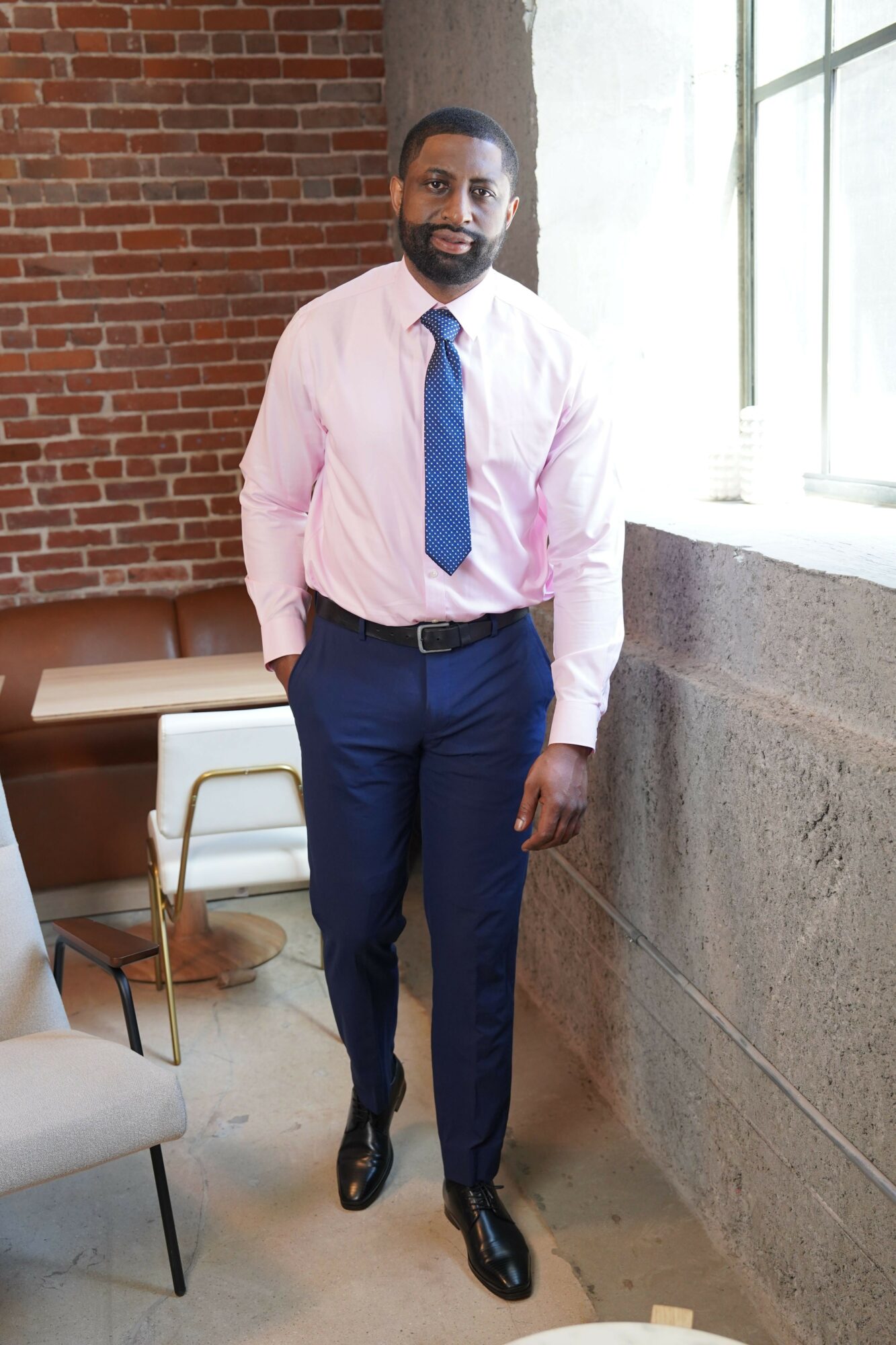 Man in pink shirt and blue tie standing indoors near a window and brick wall.