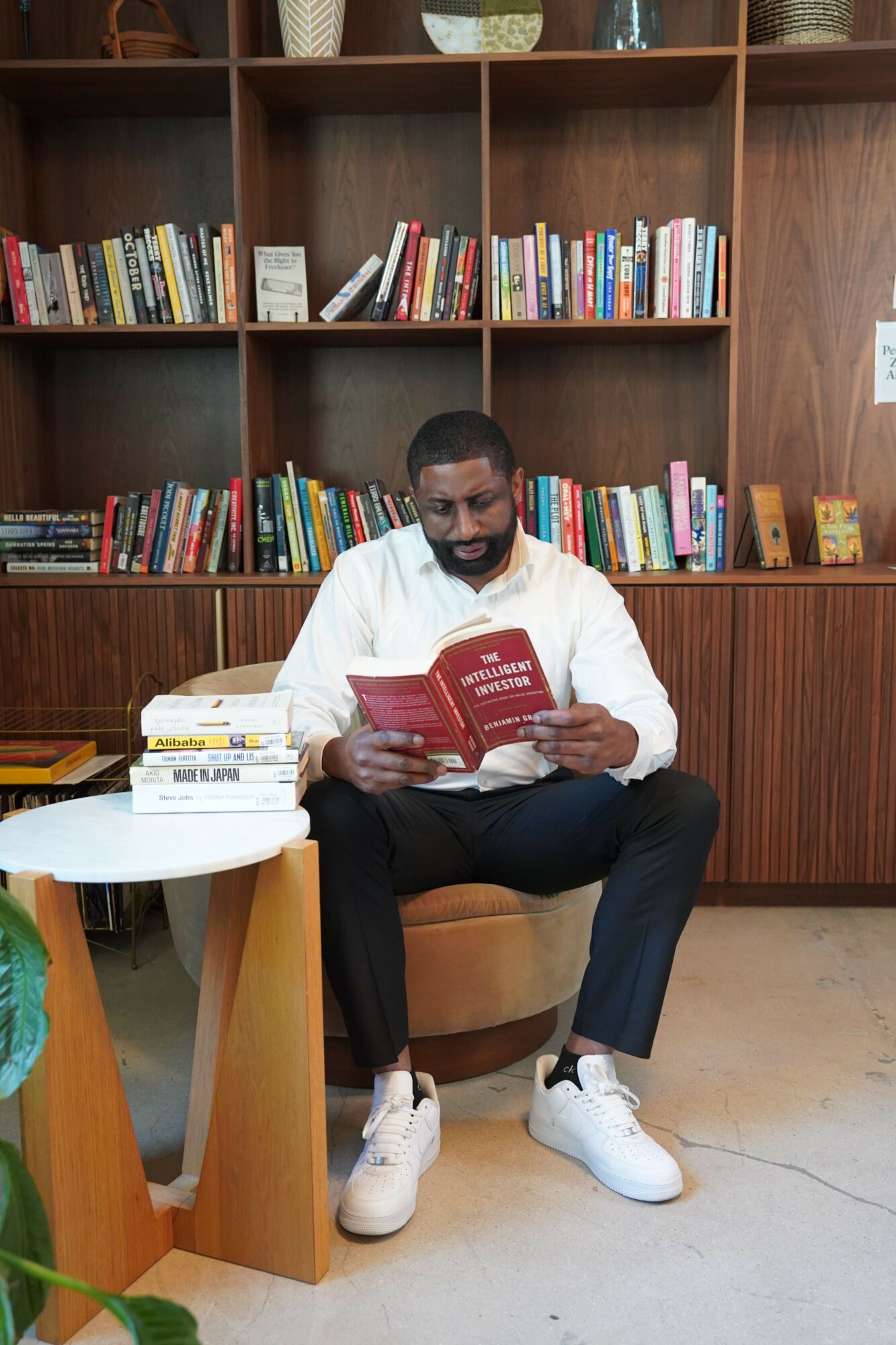 Man sitting in a chair reading a red book, bookshelf filled with books behind him, small table nearby.