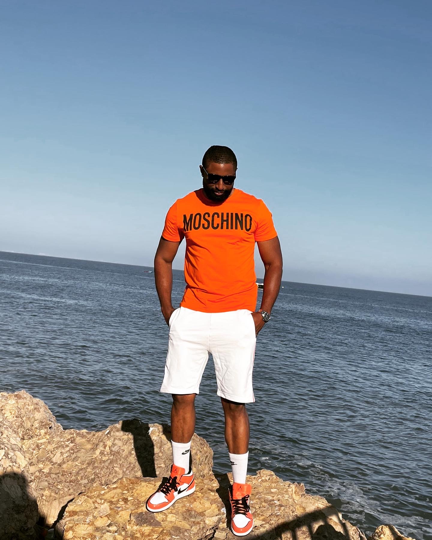 Man standing on rocks by the sea, wearing orange Moschino shirt, white shorts, and sneakers, with hands in pockets.