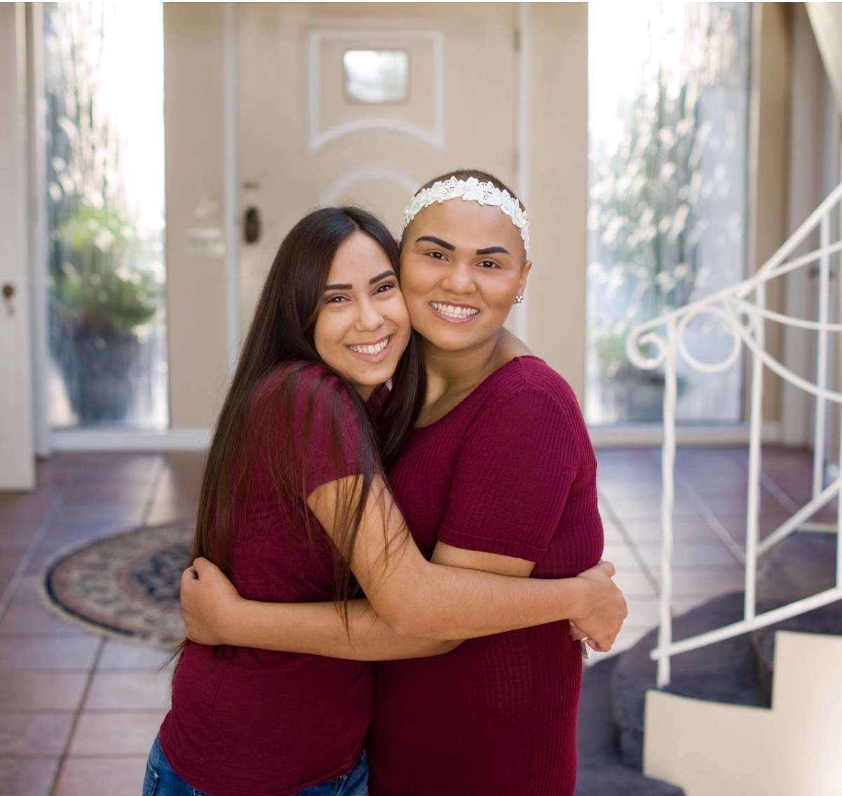 Two women hugging indoors, smiling, with a door and window in the background.