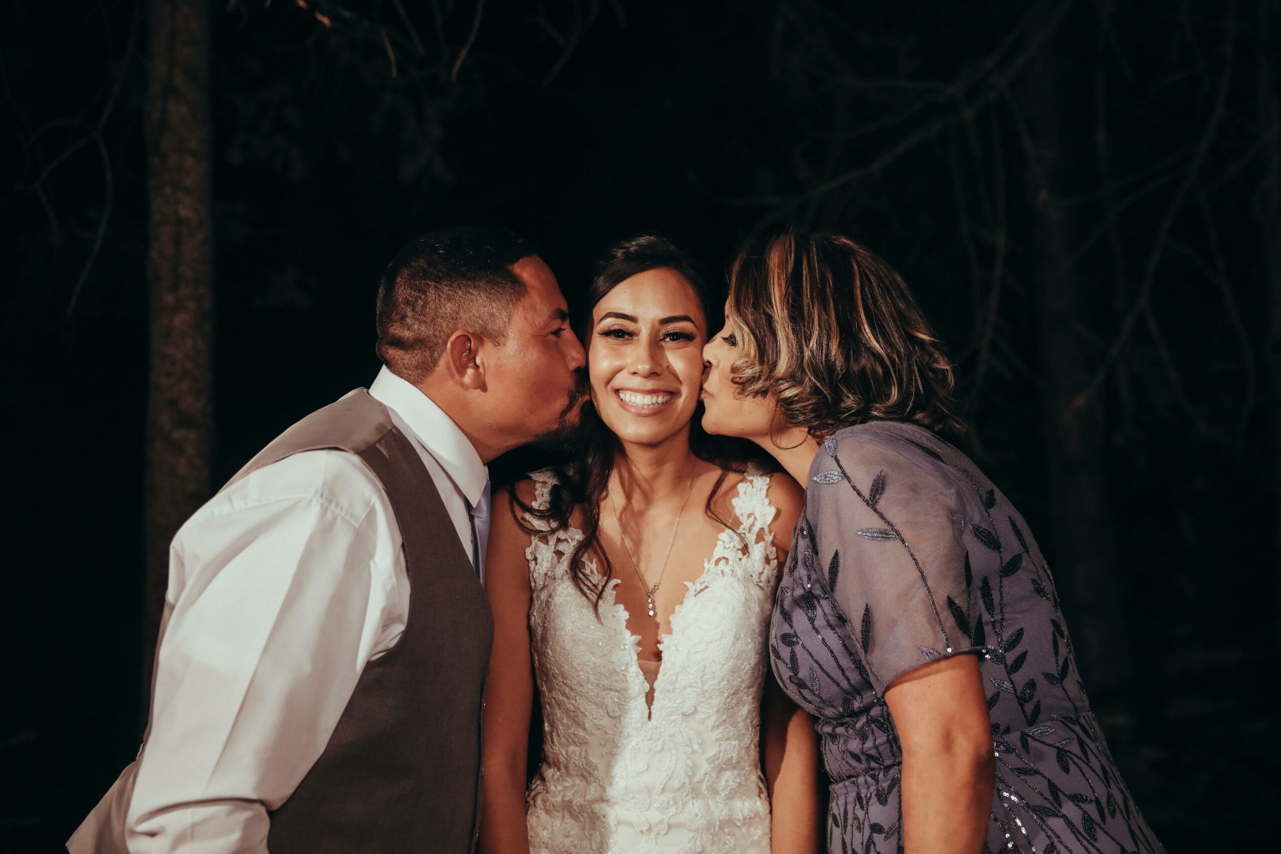 Three people outdoors at night, two women kissing a smiling woman in the middle on her cheeks.