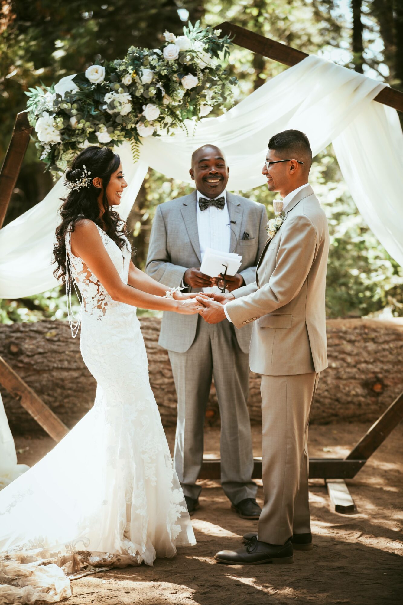 Bride and groom holding hands, officiant smiling, outdoor wedding ceremony with floral arch and trees in background.