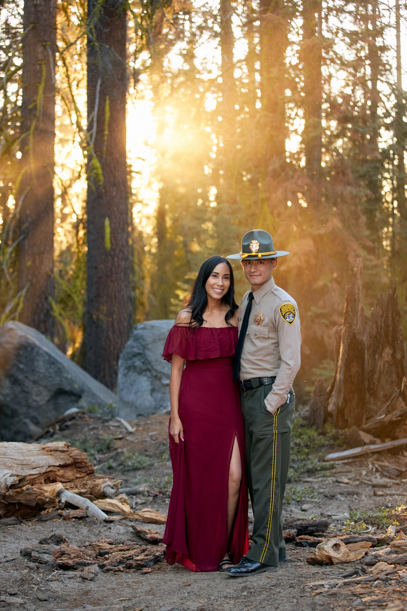 A woman in a burgundy dress and a man in a sheriff's uniform stand together in a forest with sunlight filtering through trees.
