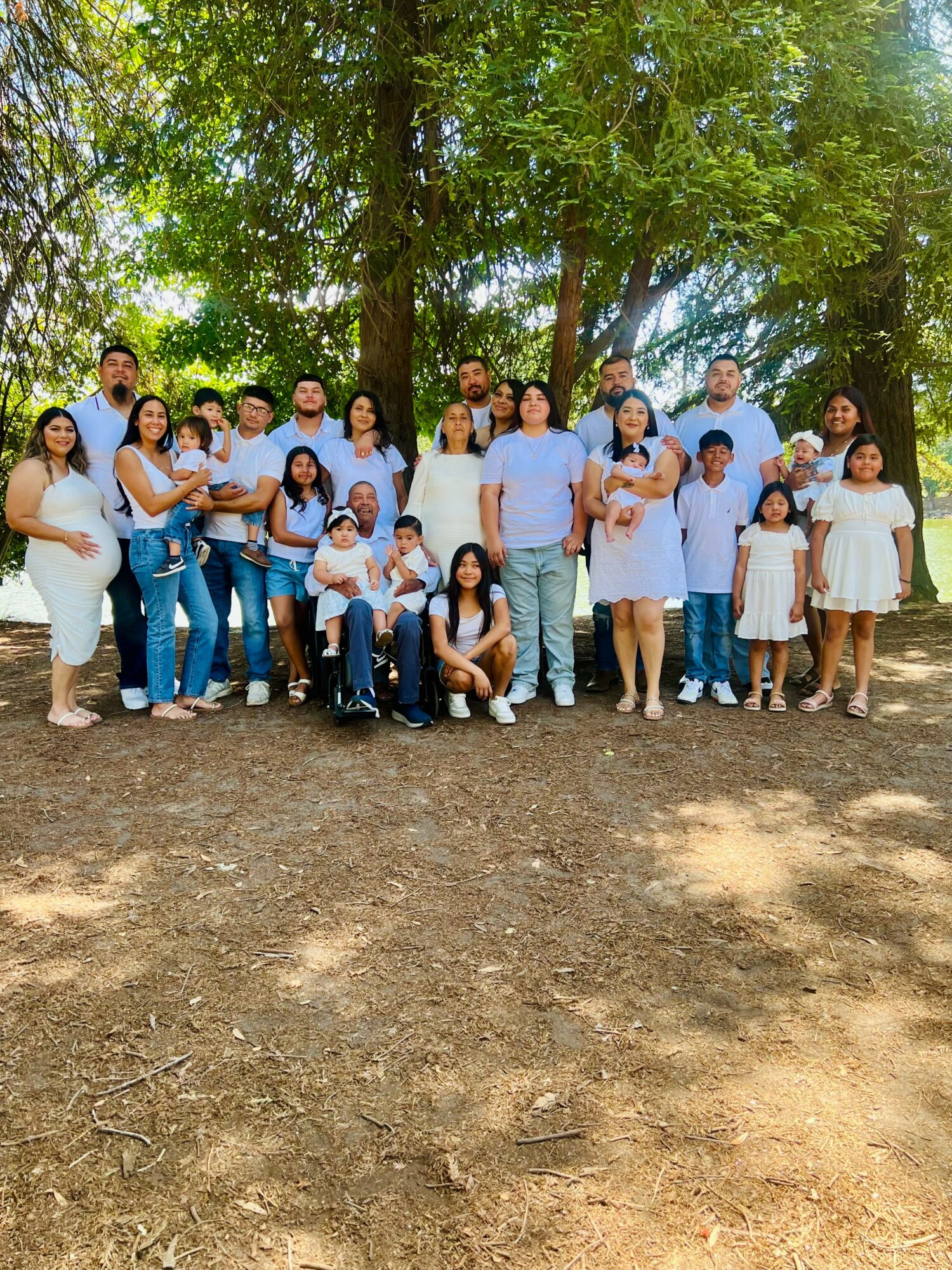 Group of people outdoors in a park with trees, standing and sitting on dirt ground, smiling at the camera.