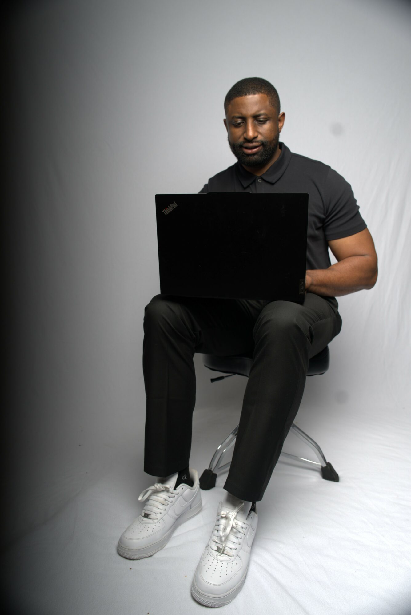 Man sitting on a chair using a laptop, wearing a black shirt and white sneakers, against a plain background.