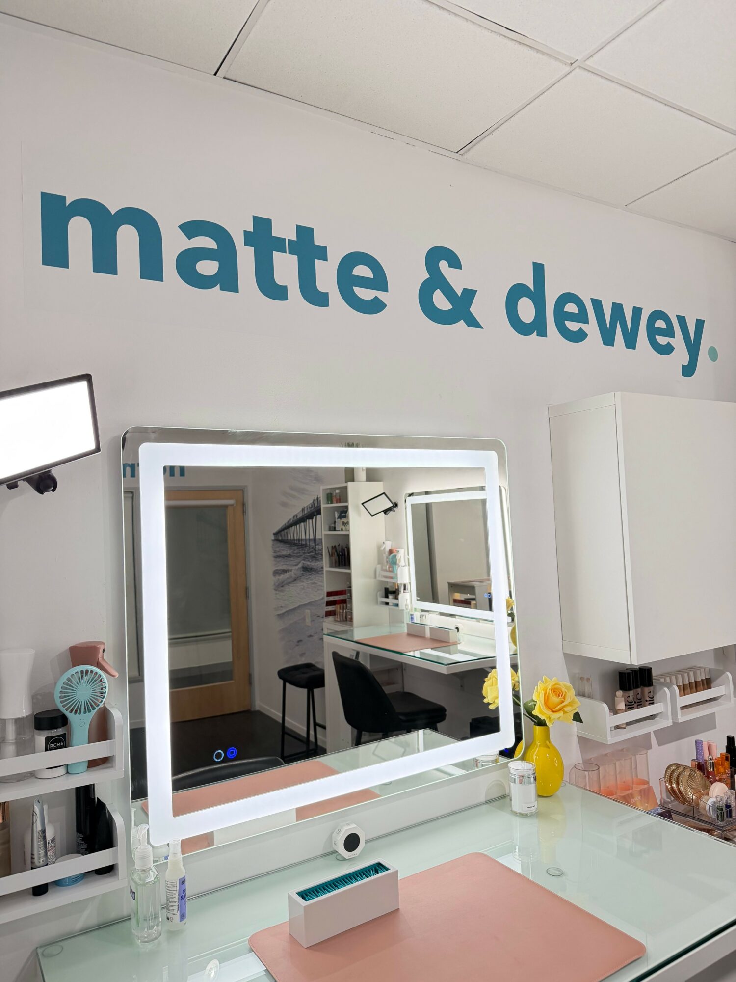 Beauty salon with a mirror, counter, and a yellow flower vase, wall sign reads 'matte & dewey,' ceiling tiles visible.