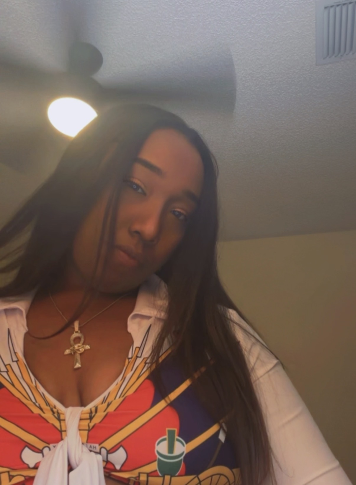 Woman with long dark hair wearing a white shirt and gold necklace, indoors with ceiling light visible.