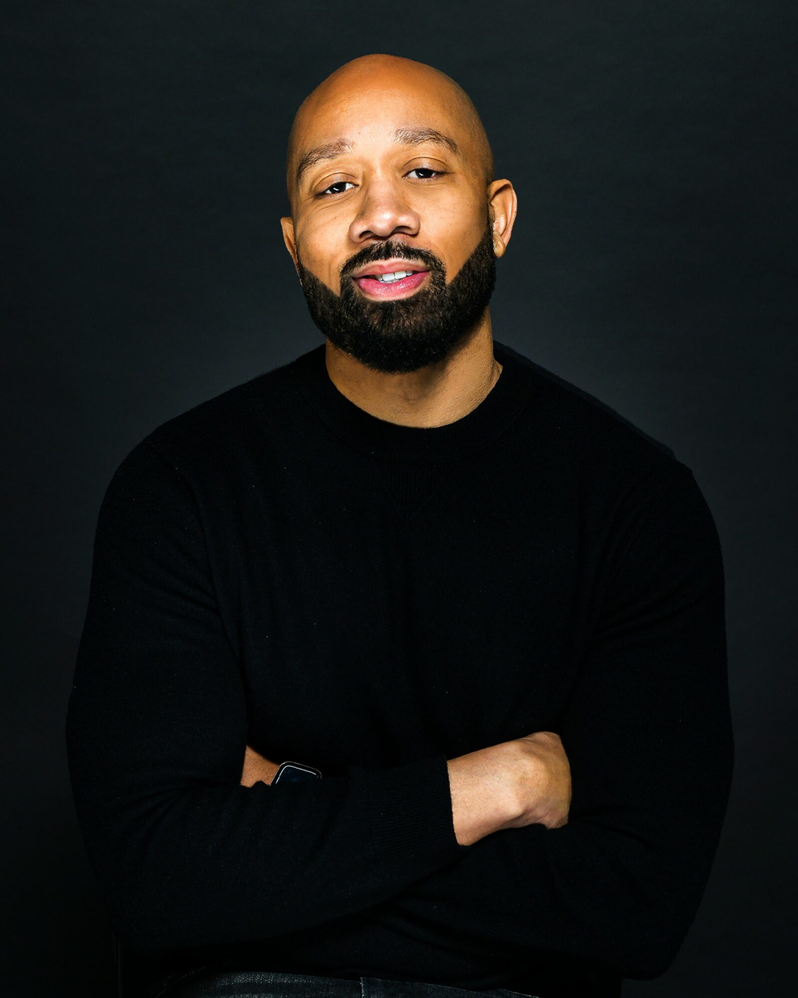 Man with a beard and bald head wearing a black long-sleeve shirt, crossing arms, against dark background.
