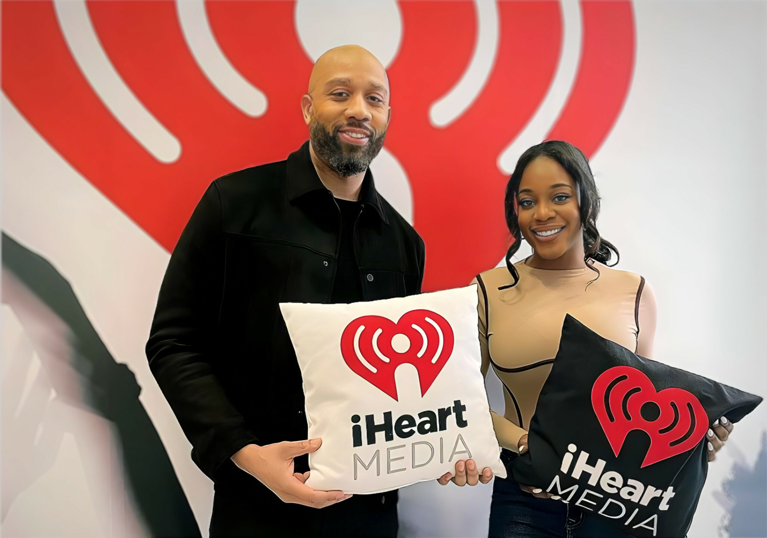 Two people holding iHeart Media signs, smiling, standing in front of a red heart logo backdrop.