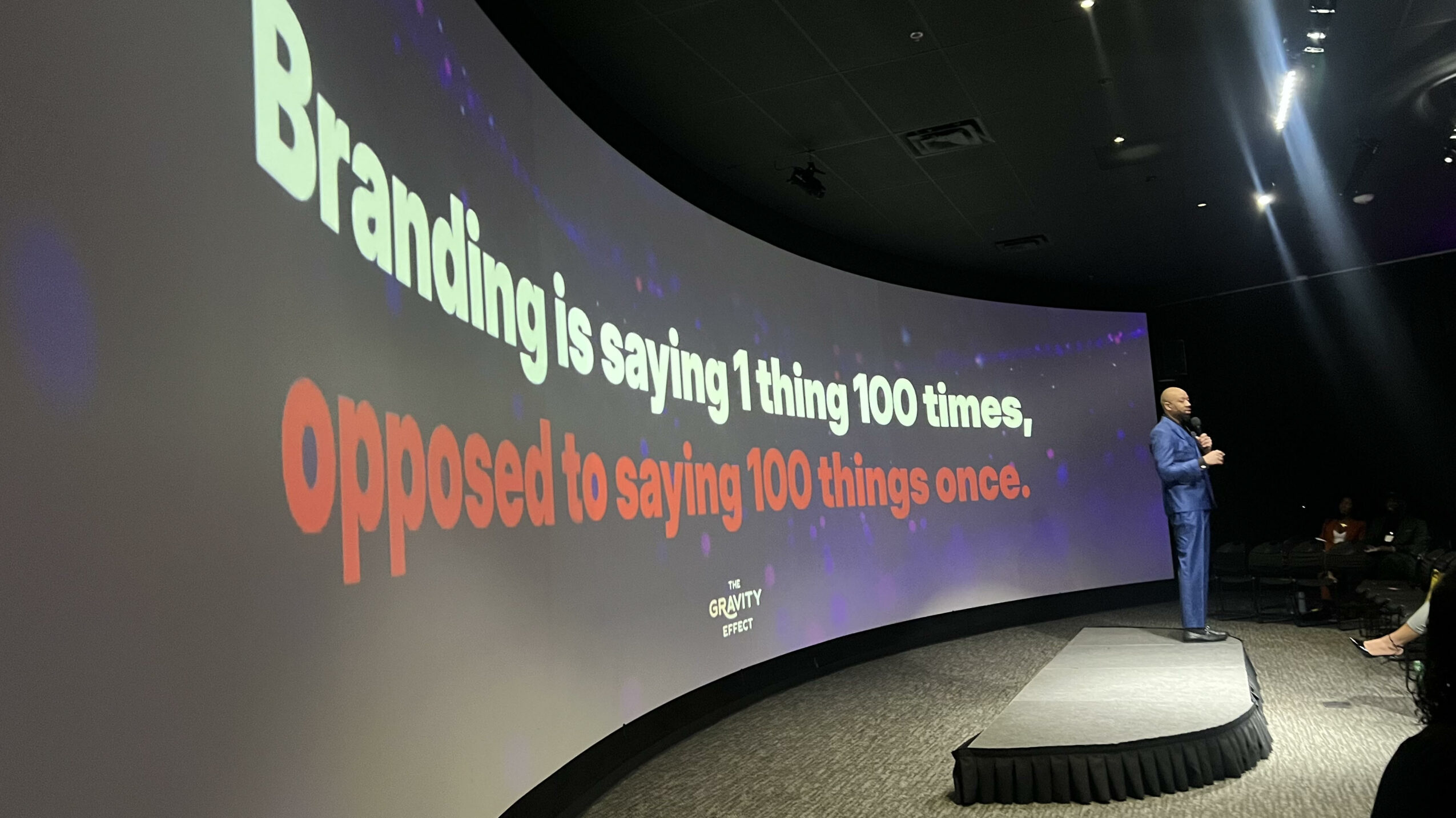 Person standing on stage in front of a large curved screen with text about branding and opposition.
