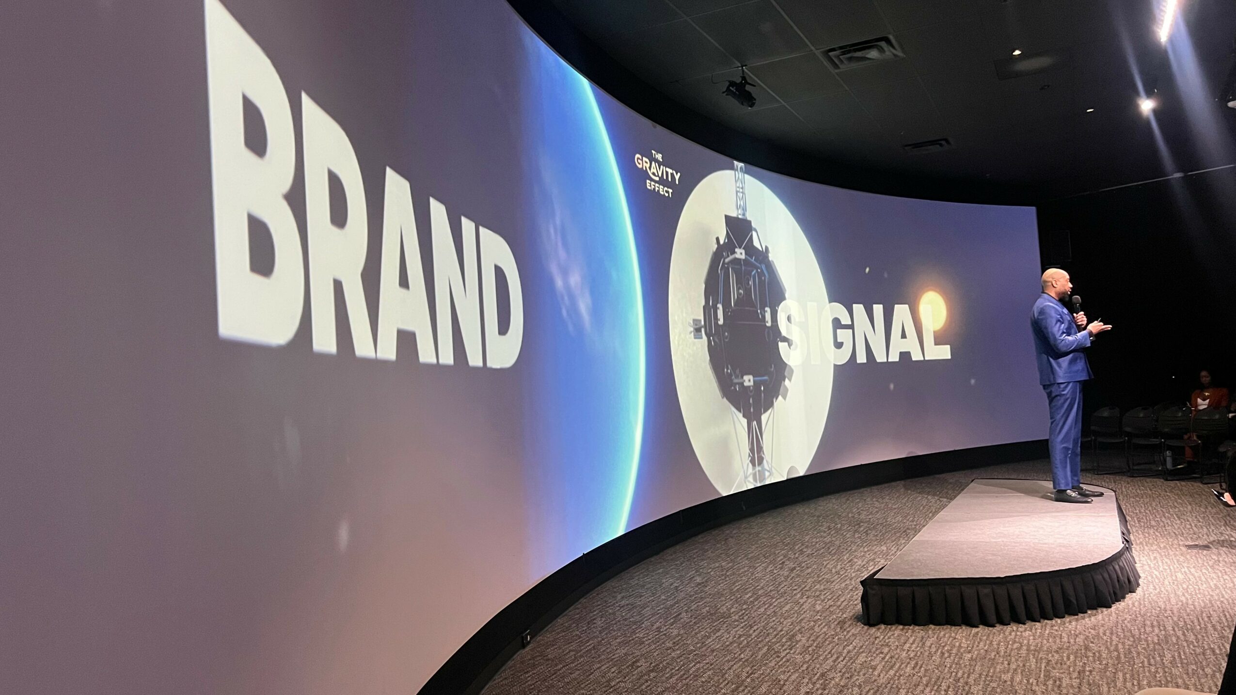 A speaker stands on a stage in front of a large screen displaying the words 'BRAND' and 'SIGNAL' with a circular graphic.