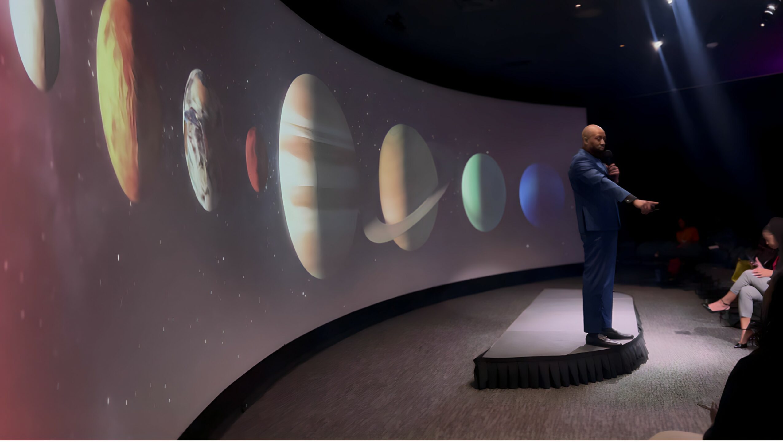 A man in a suit stands on a platform in front of a curved screen displaying planets and celestial objects.