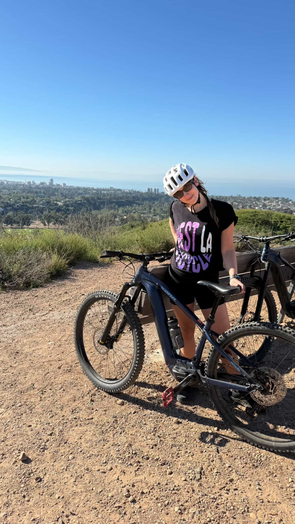 Person wearing a helmet and sunglasses standing next to a bicycle on a dirt trail with a landscape and blue sky in the background.