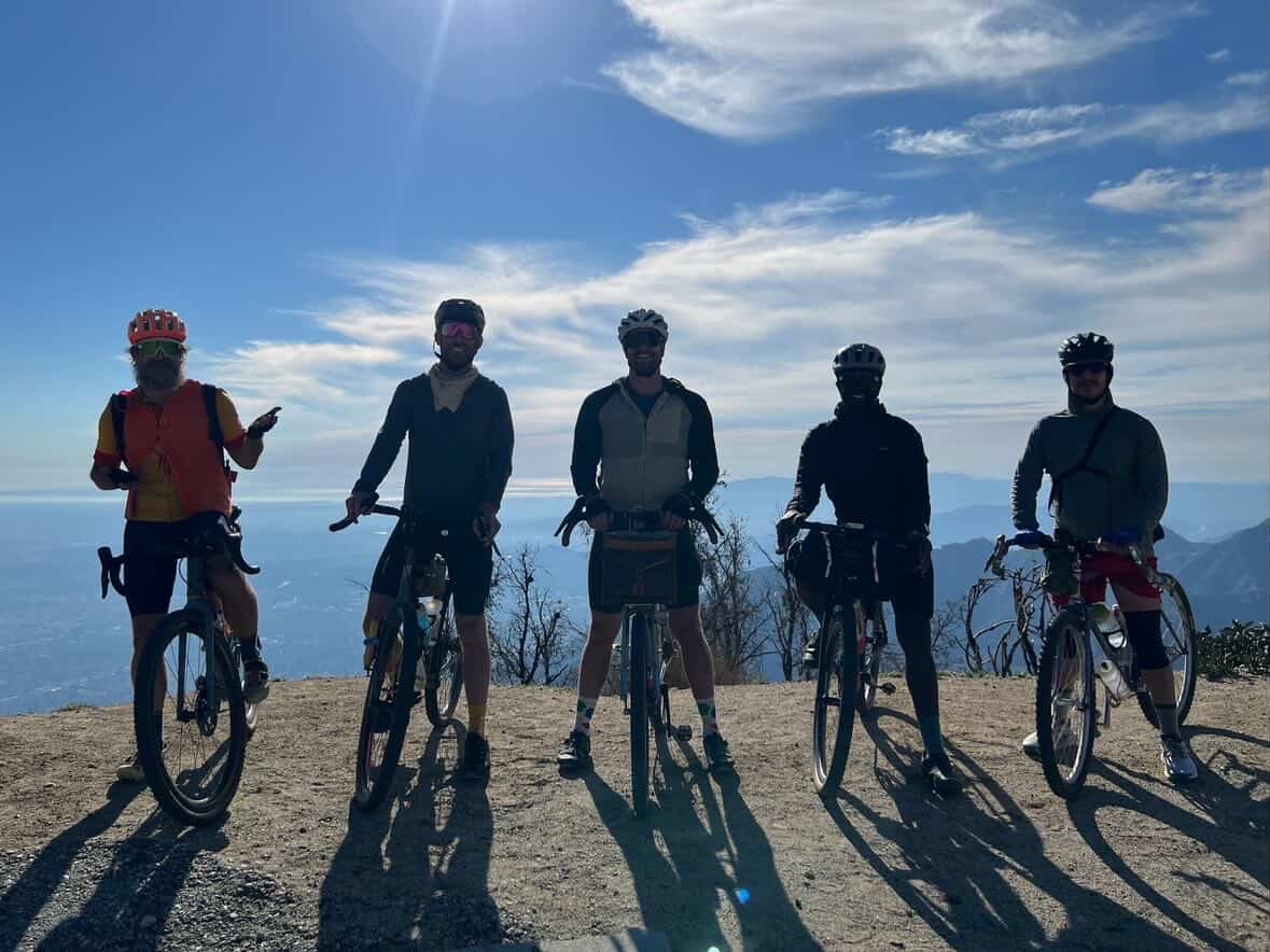 Five people with bicycles stand on a hilltop with a mountain view and blue sky in the background.