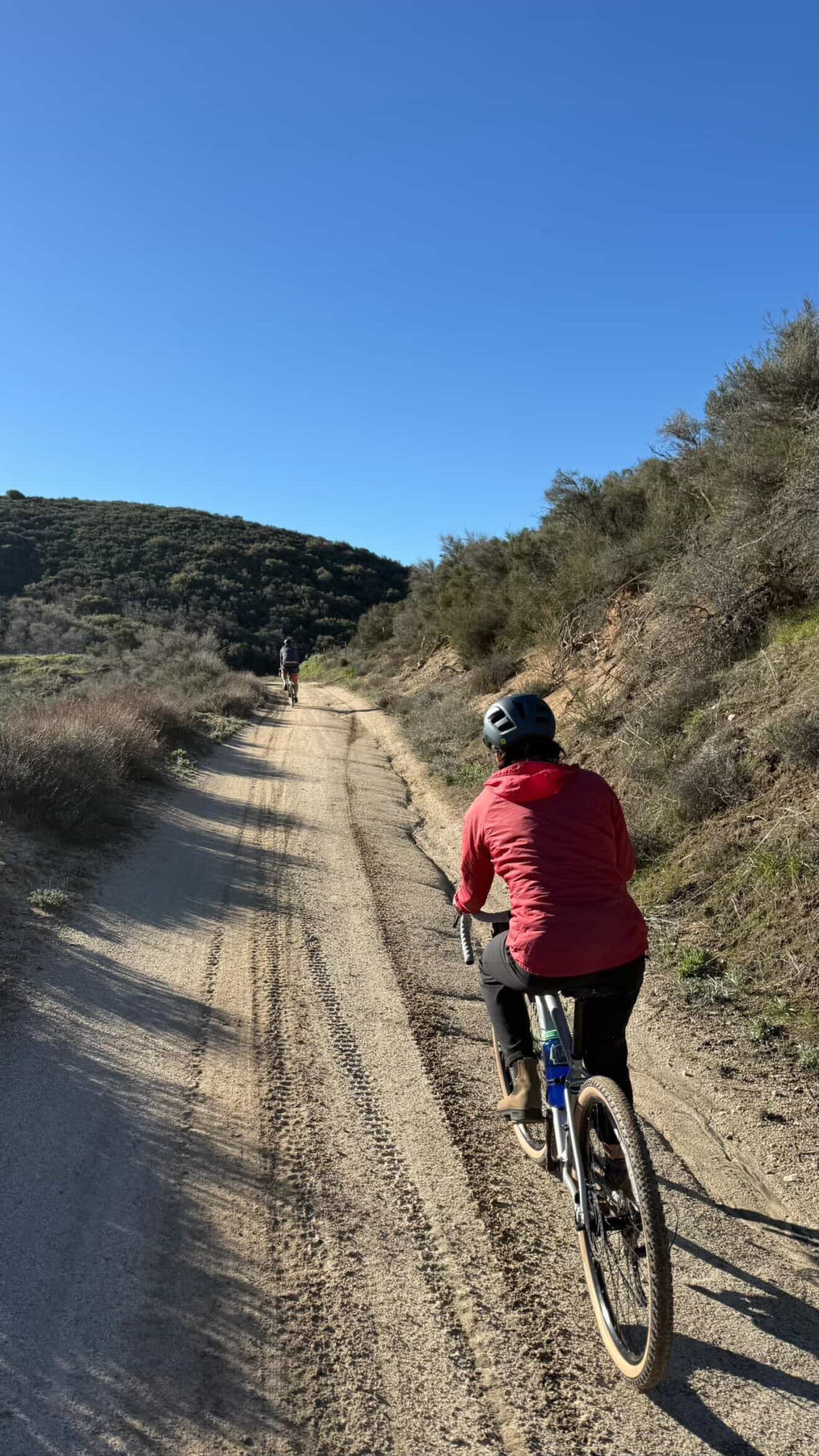 Person riding a bicycle on a dirt trail surrounded by hills and bushes under a clear blue sky.