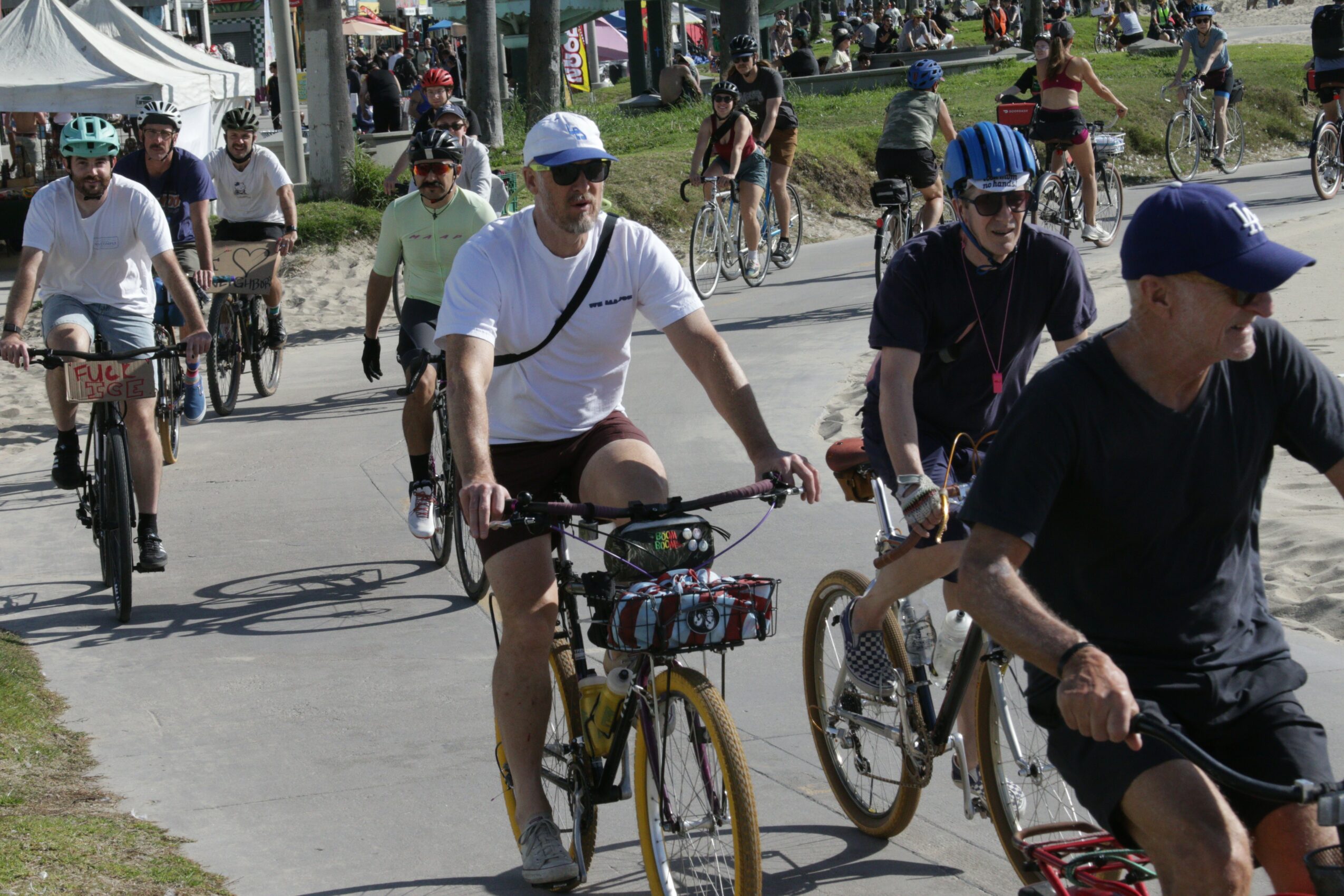 Group of people riding bicycles on a paved path outdoors, some wearing helmets and sunglasses, with trees and tents in background.