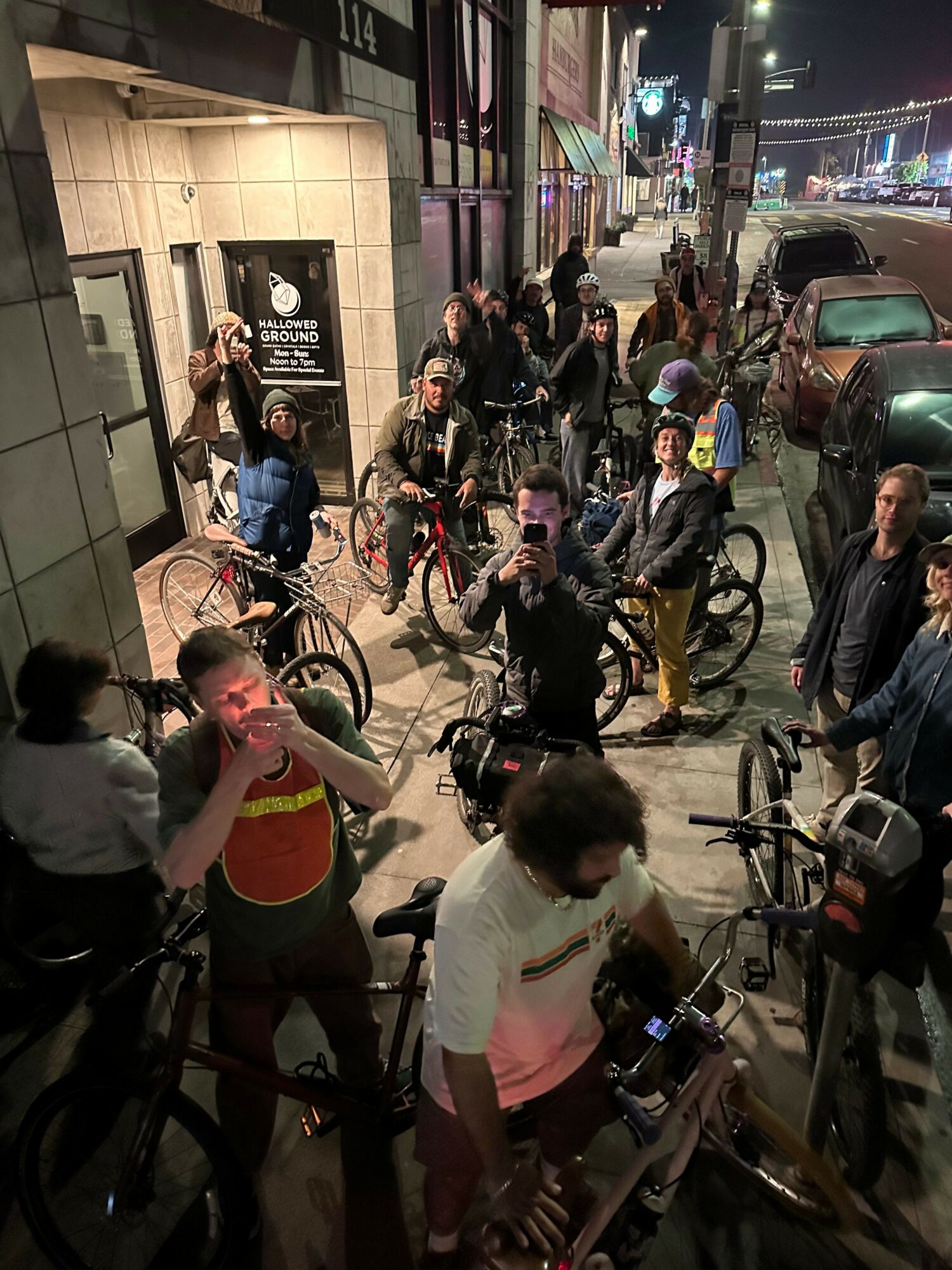 Group of people with bicycles gathered on a city sidewalk at night, illuminated by streetlights and building lights.