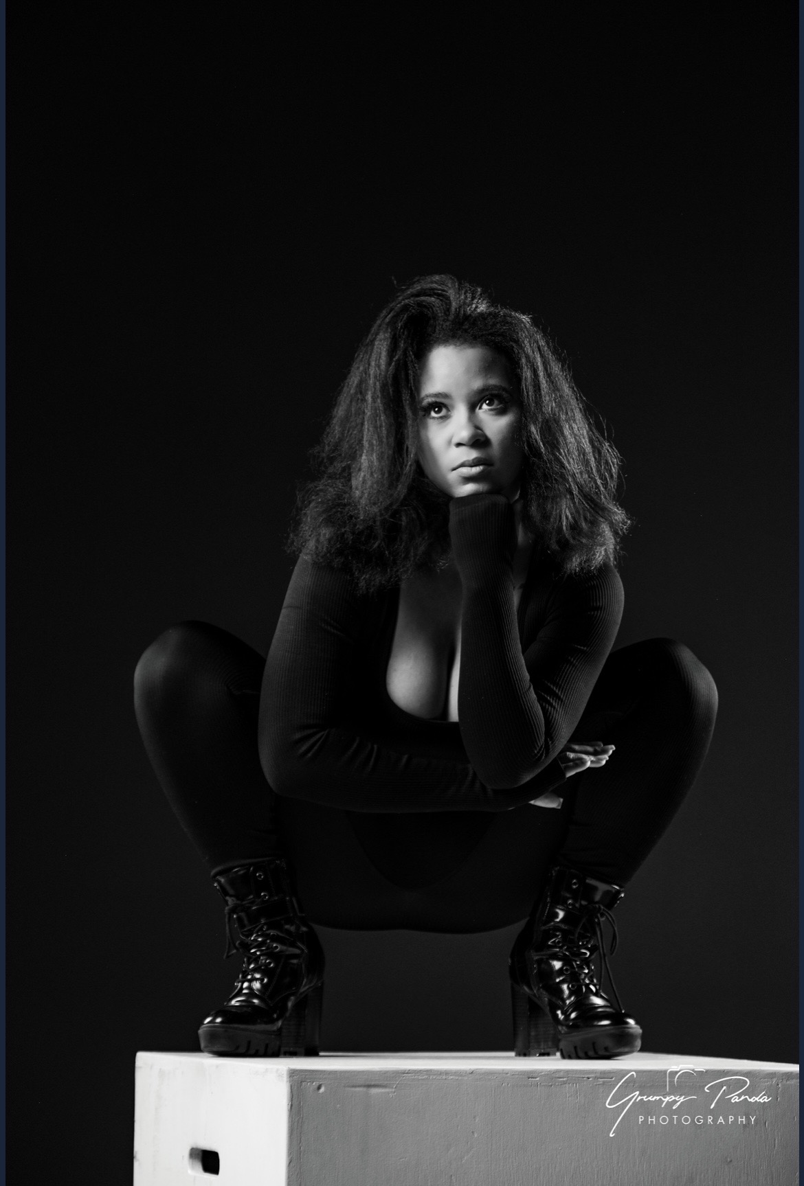 Woman crouching on a white box, looking at camera, with dark background, wearing black outfit and boots, hair voluminous.