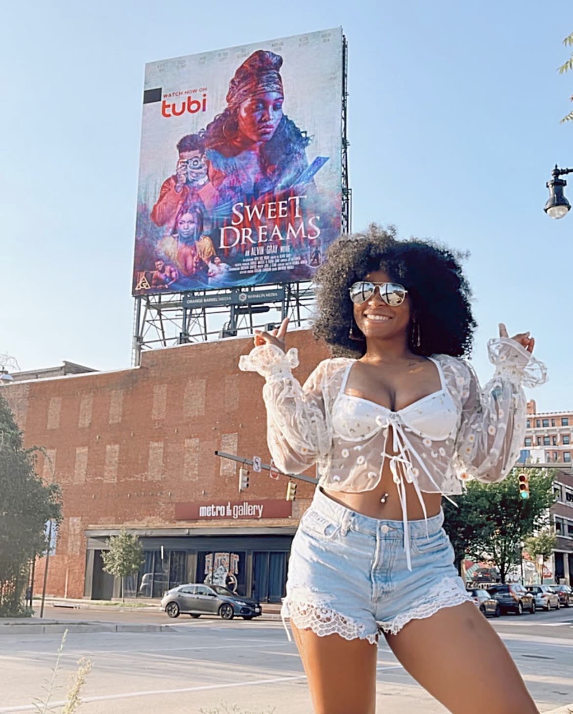 Young woman with curly hair and glasses smiling outdoors, wearing a white lace crop top and denim shorts.