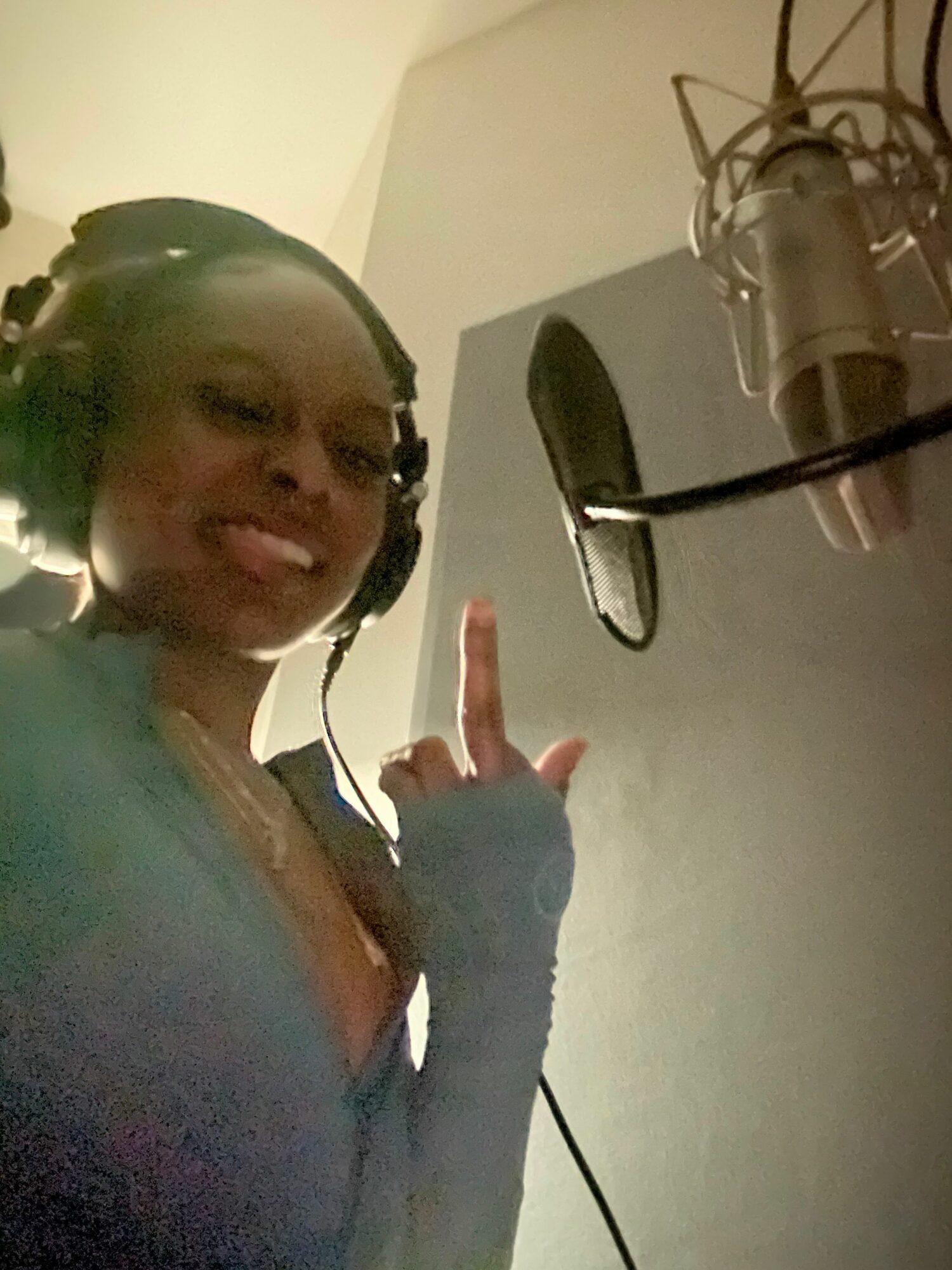 Woman in a recording studio with headphones, standing near a microphone and pop filter, smiling and making a hand gesture.