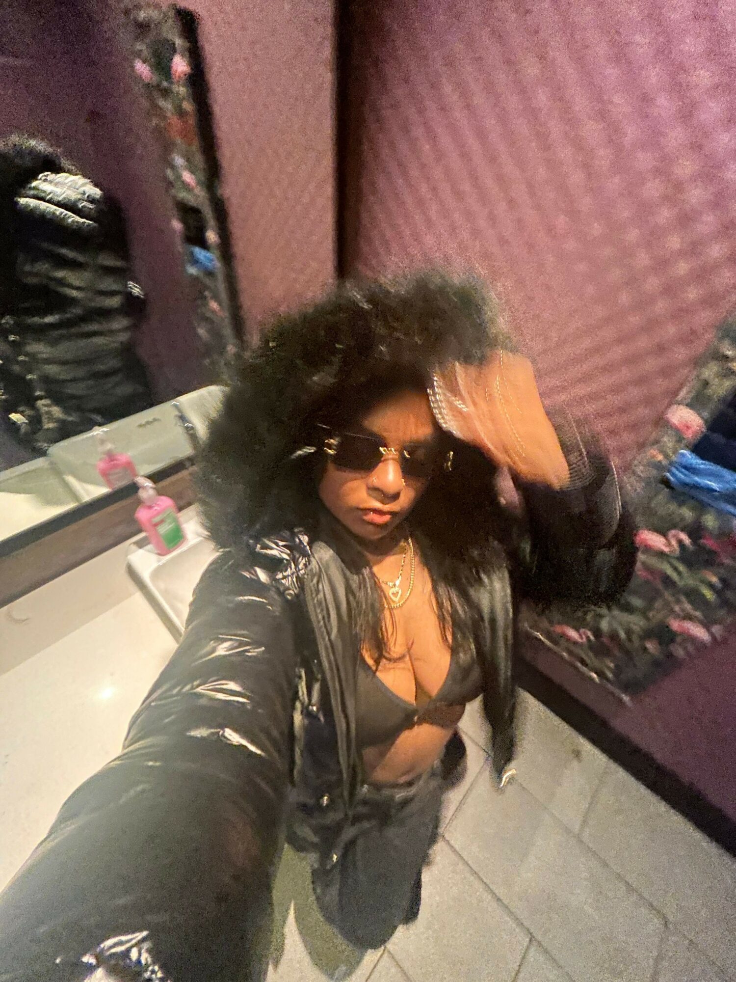 Woman with curly hair wearing sunglasses and a leather jacket, taking a selfie indoors.