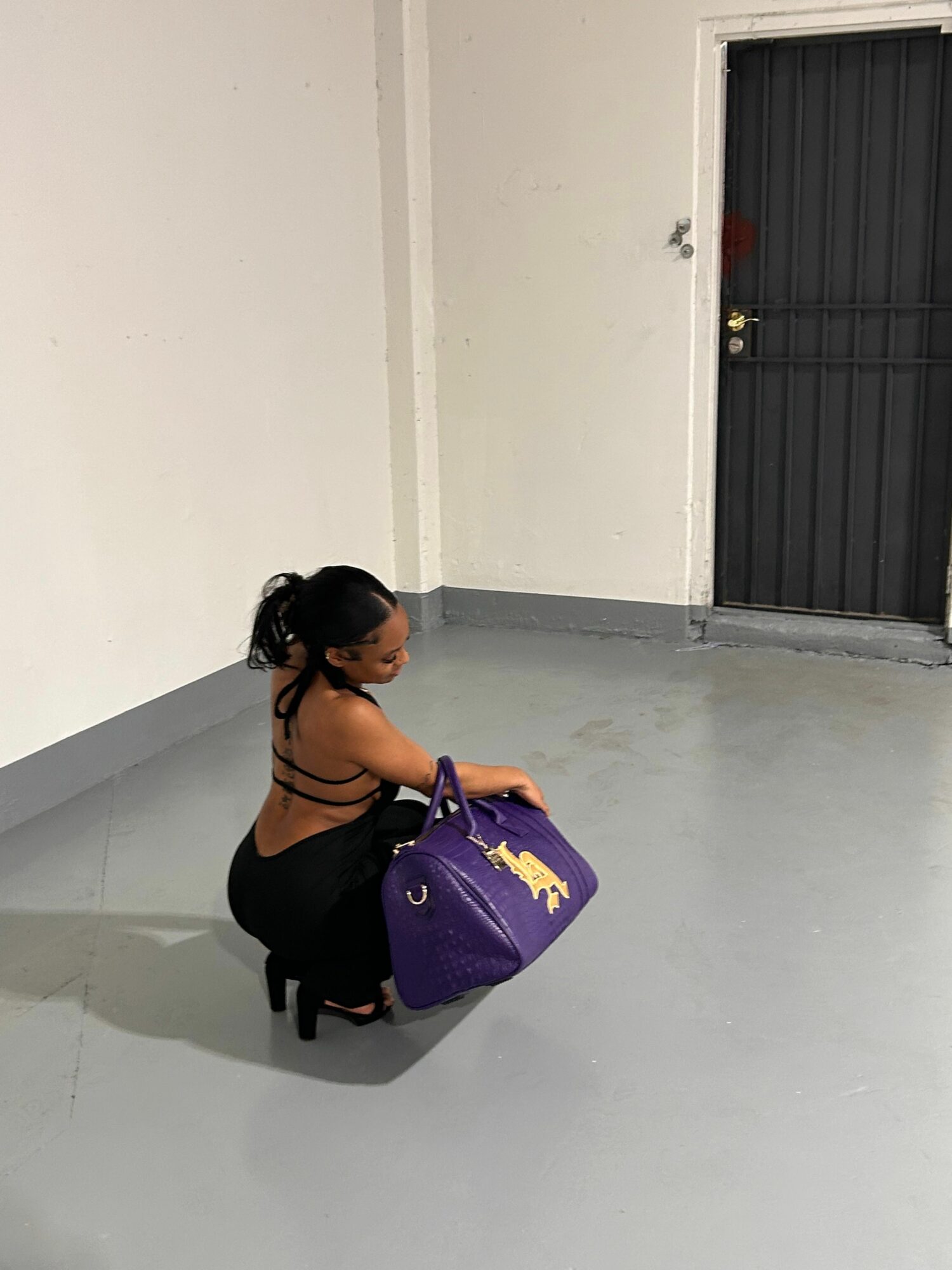Woman crouching indoors, holding a purple bag, wearing black clothing and high heels, near a door.