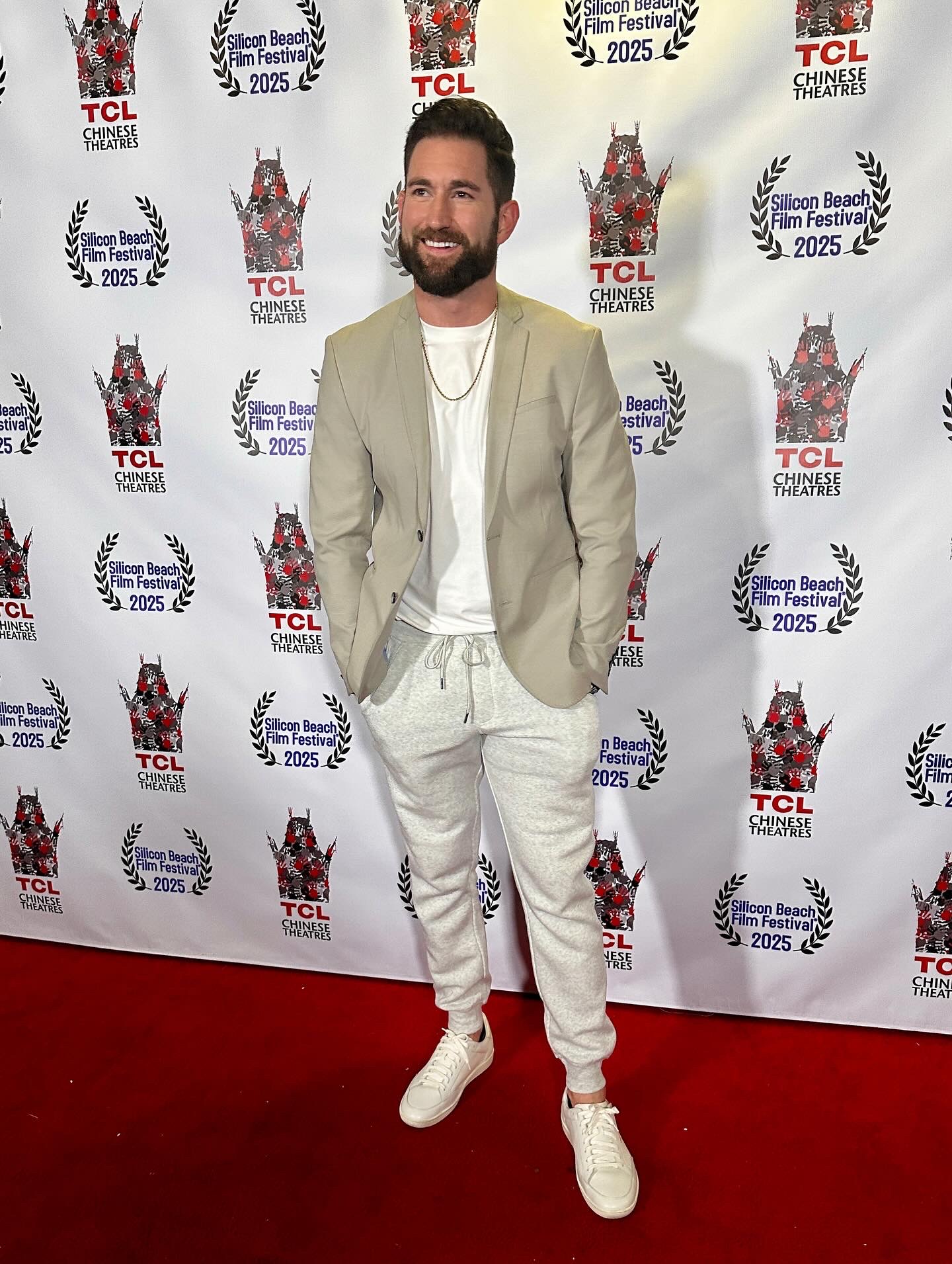 Man with beard in beige blazer and white pants standing on red carpet in front of event backdrop.