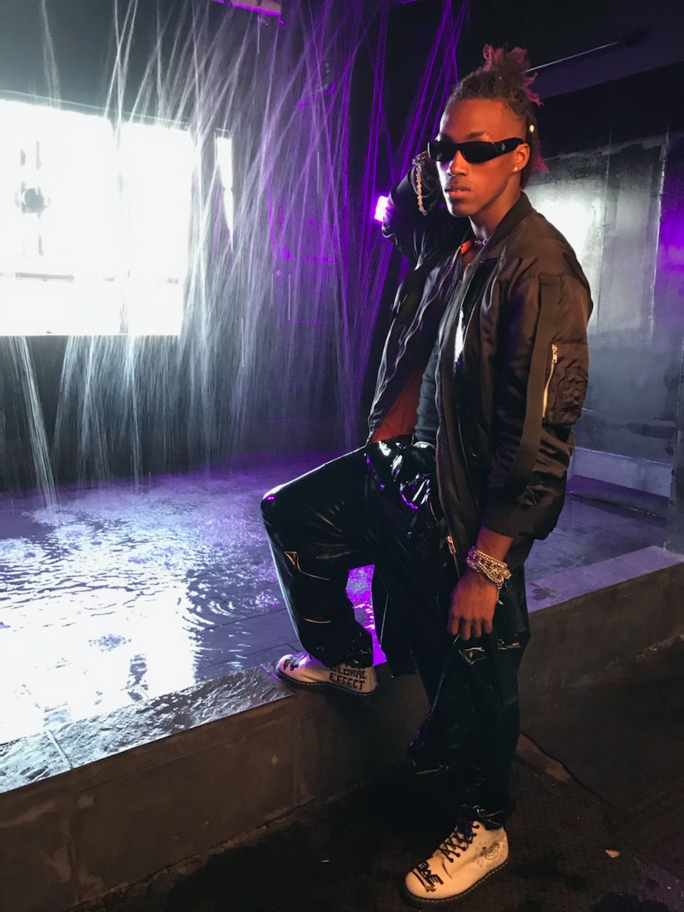 Person with sunglasses and braided hair standing near water feature with purple lighting, wearing a black jacket and sneakers.