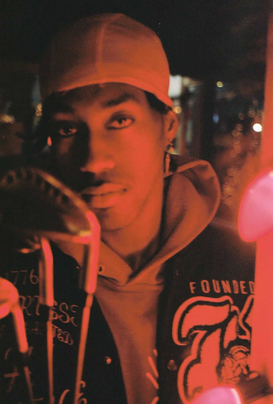 Young person wearing a cap and hoodie, illuminated by pink and orange lights in a dark setting.