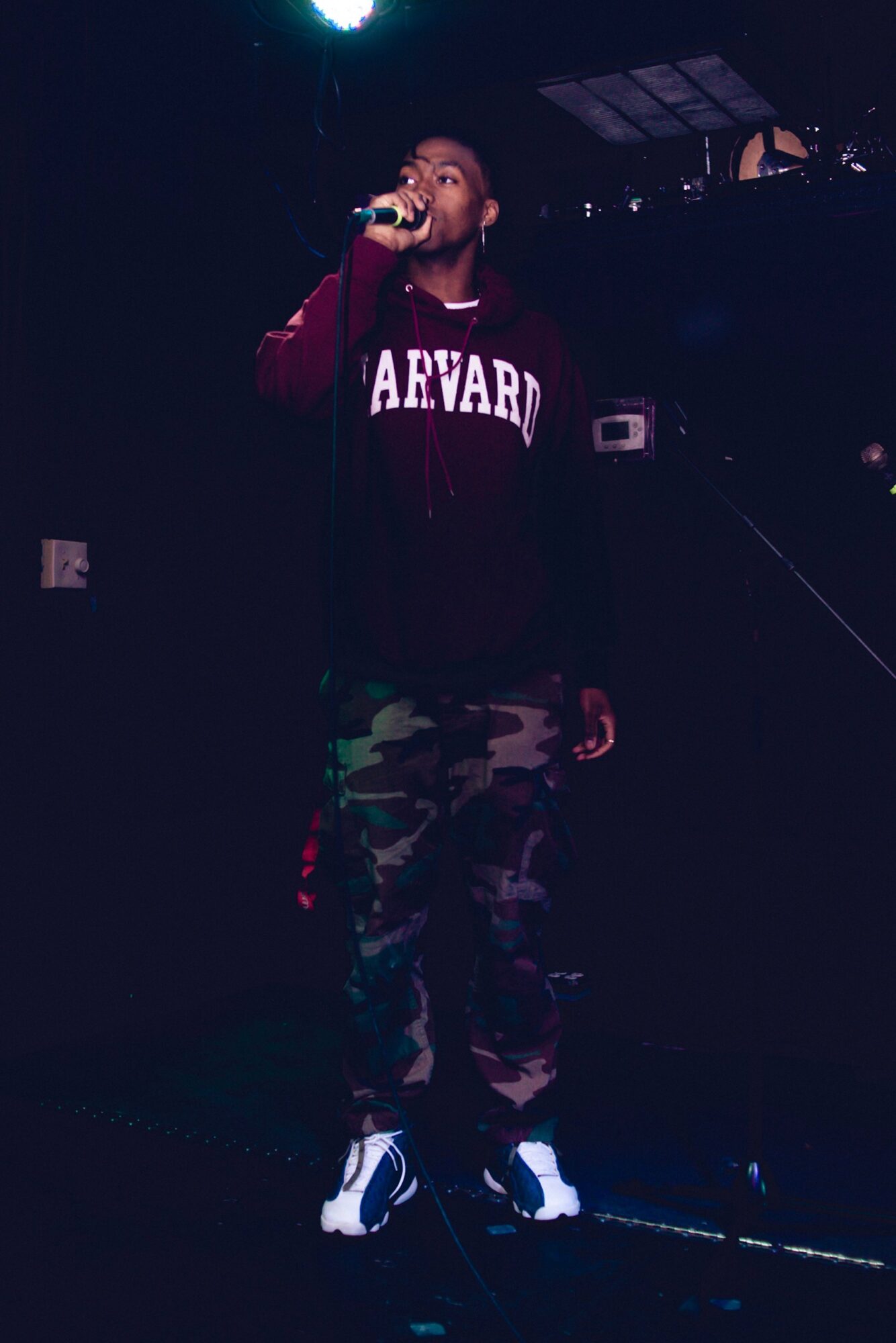 Person holding microphone on stage, wearing a Harvard jacket and camouflage pants, in a dark setting.