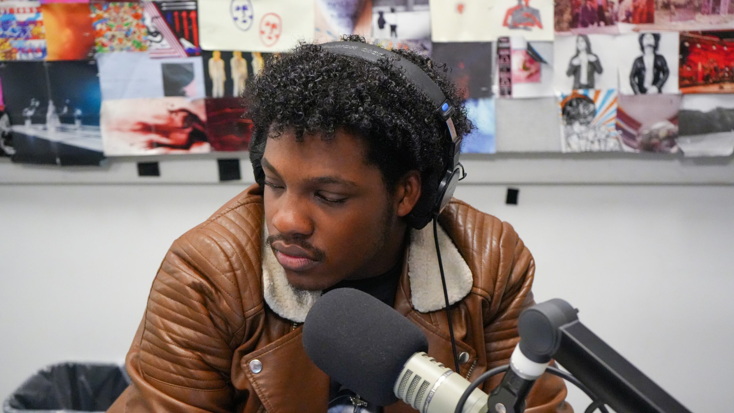 Young man with curly hair wearing headphones, speaking into a microphone in a recording studio.