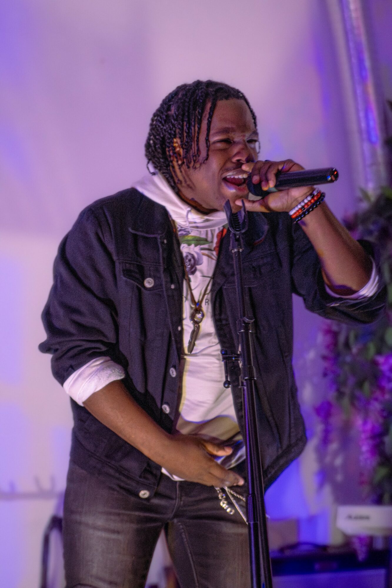 Person singing into microphone on stage with purple lighting and floral arrangement in background.