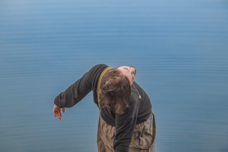 Person leaning back with head tilted, arms extended, near a calm body of water.