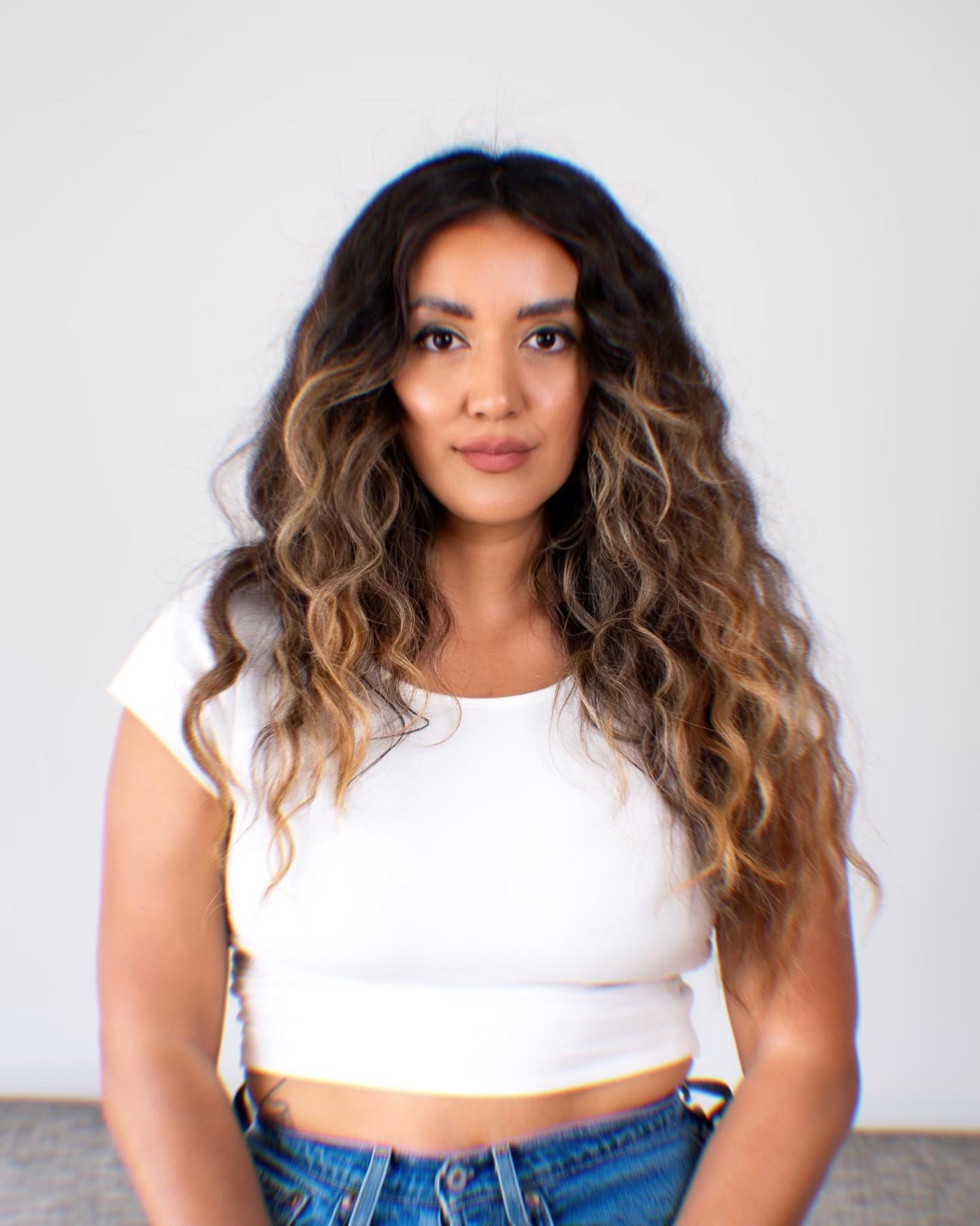 Woman with long, curly hair wearing a white crop top and blue jeans, standing against a plain background.