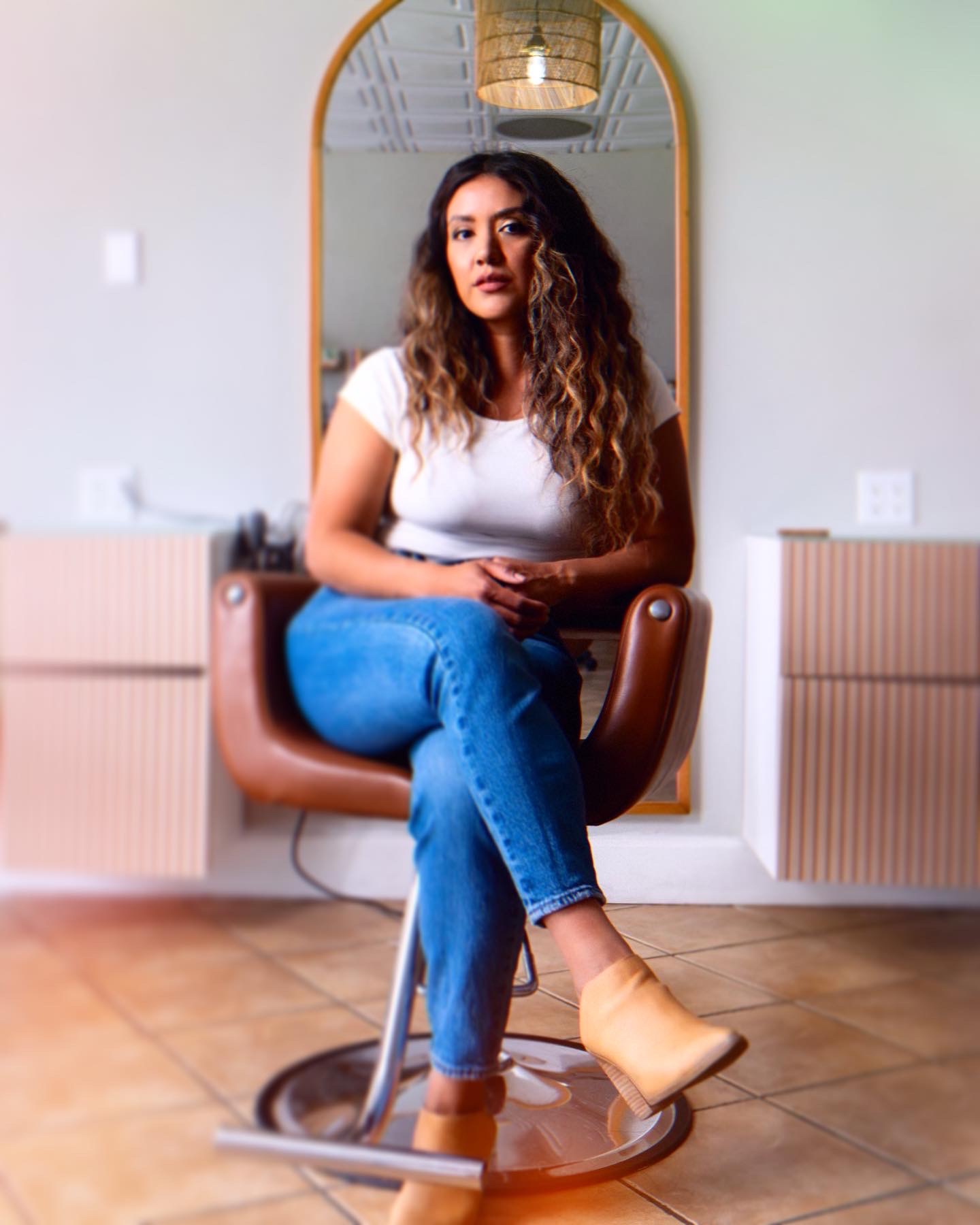 Woman with curly hair sitting on a chair in front of a mirror, wearing a white top and blue jeans.