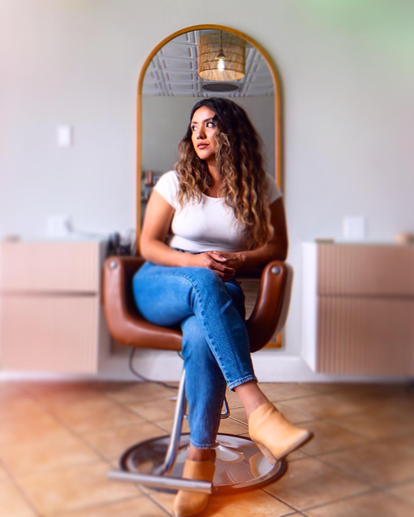 Woman with curly hair sitting on a brown chair in front of a mirror, wearing a white shirt and blue jeans.