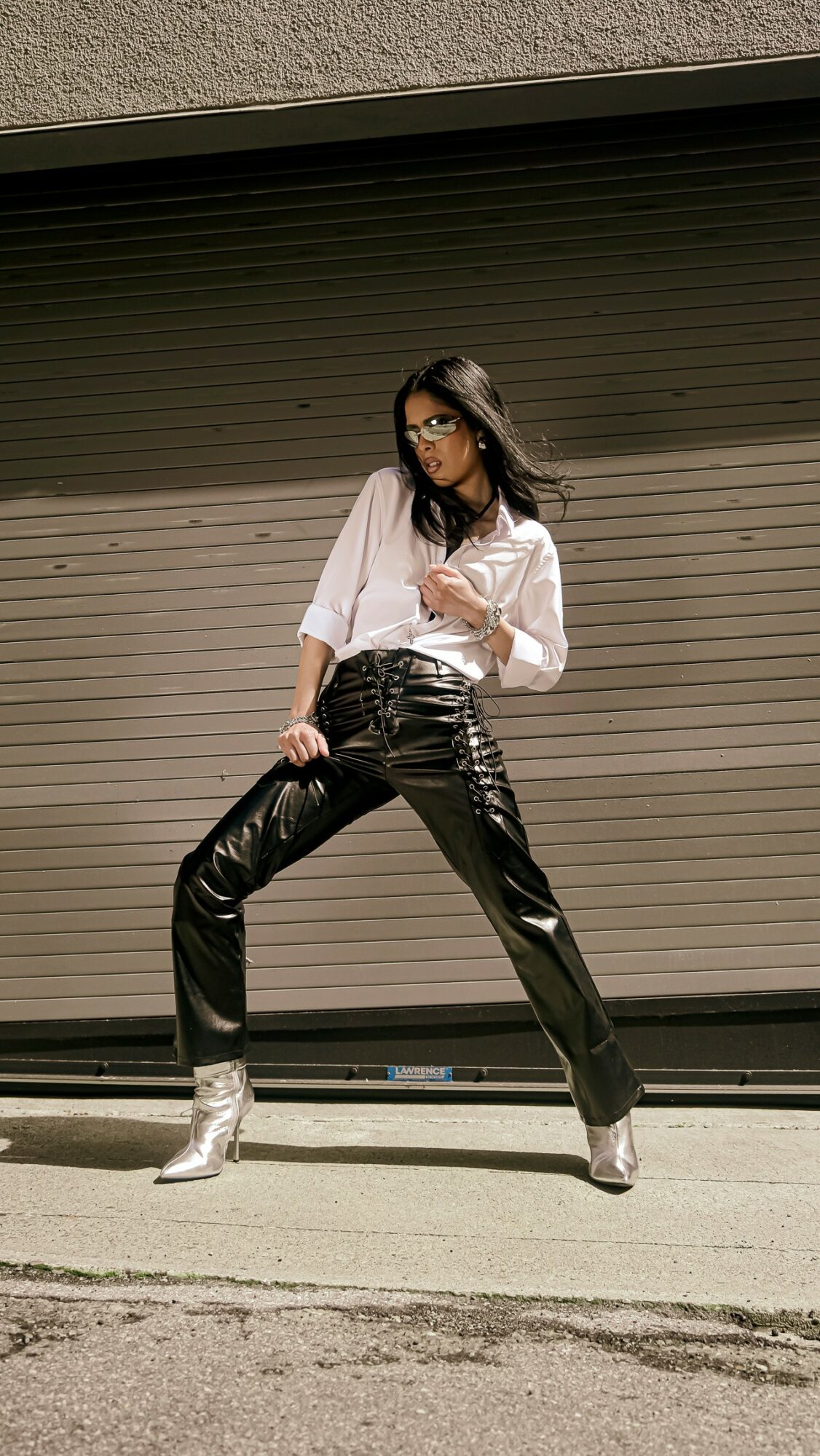 Woman in white shirt and black shiny pants stands with legs apart in front of a garage door.