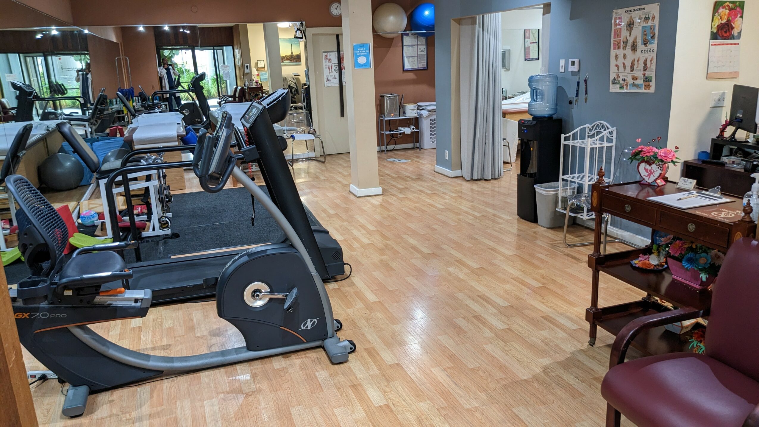 Gym with exercise machines, wooden floor, and a sitting area with chairs and a table, bright lighting.
