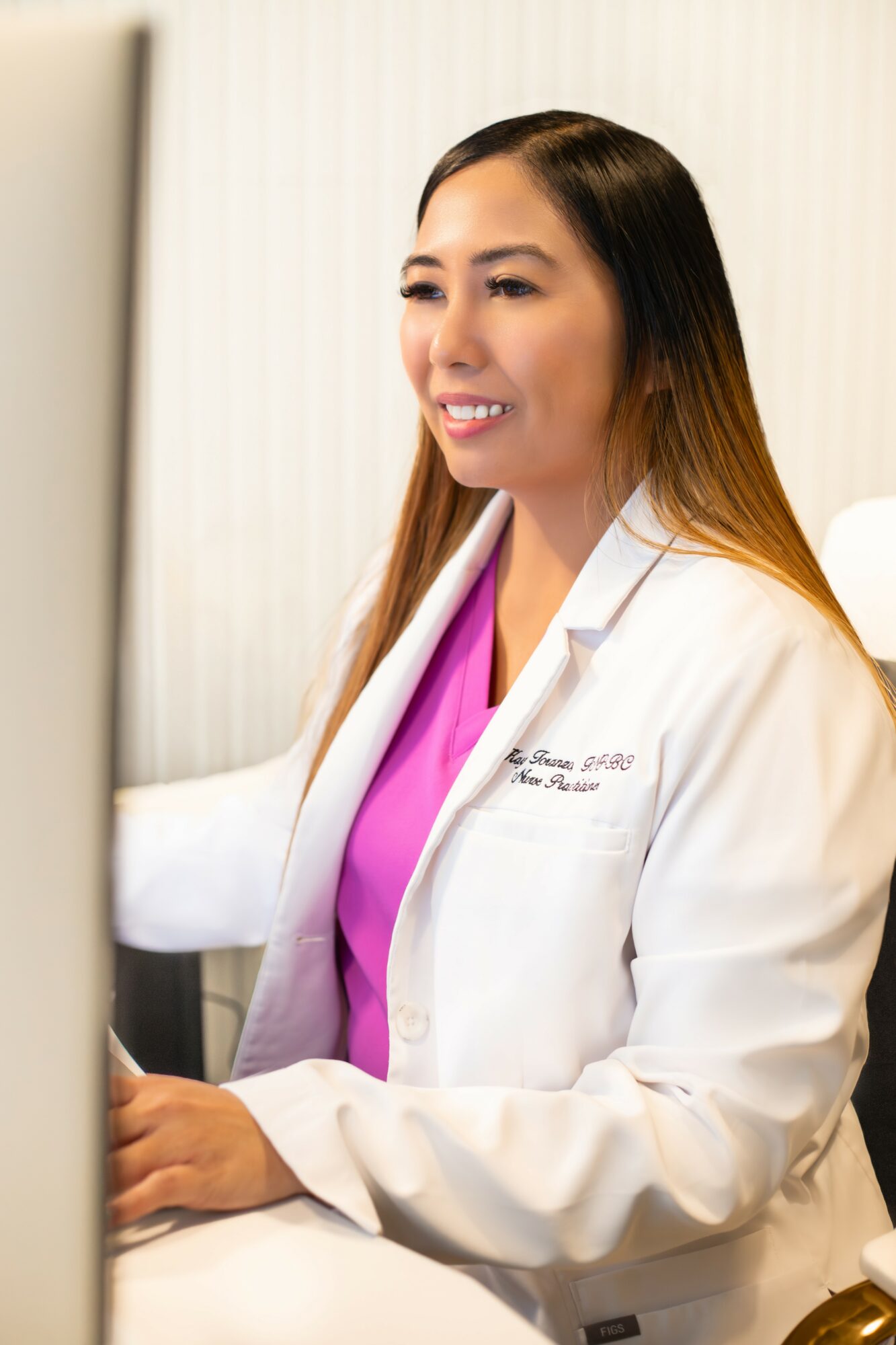 Female healthcare professional in white coat and pink scrubs working at computer, smiling, in clinical setting.