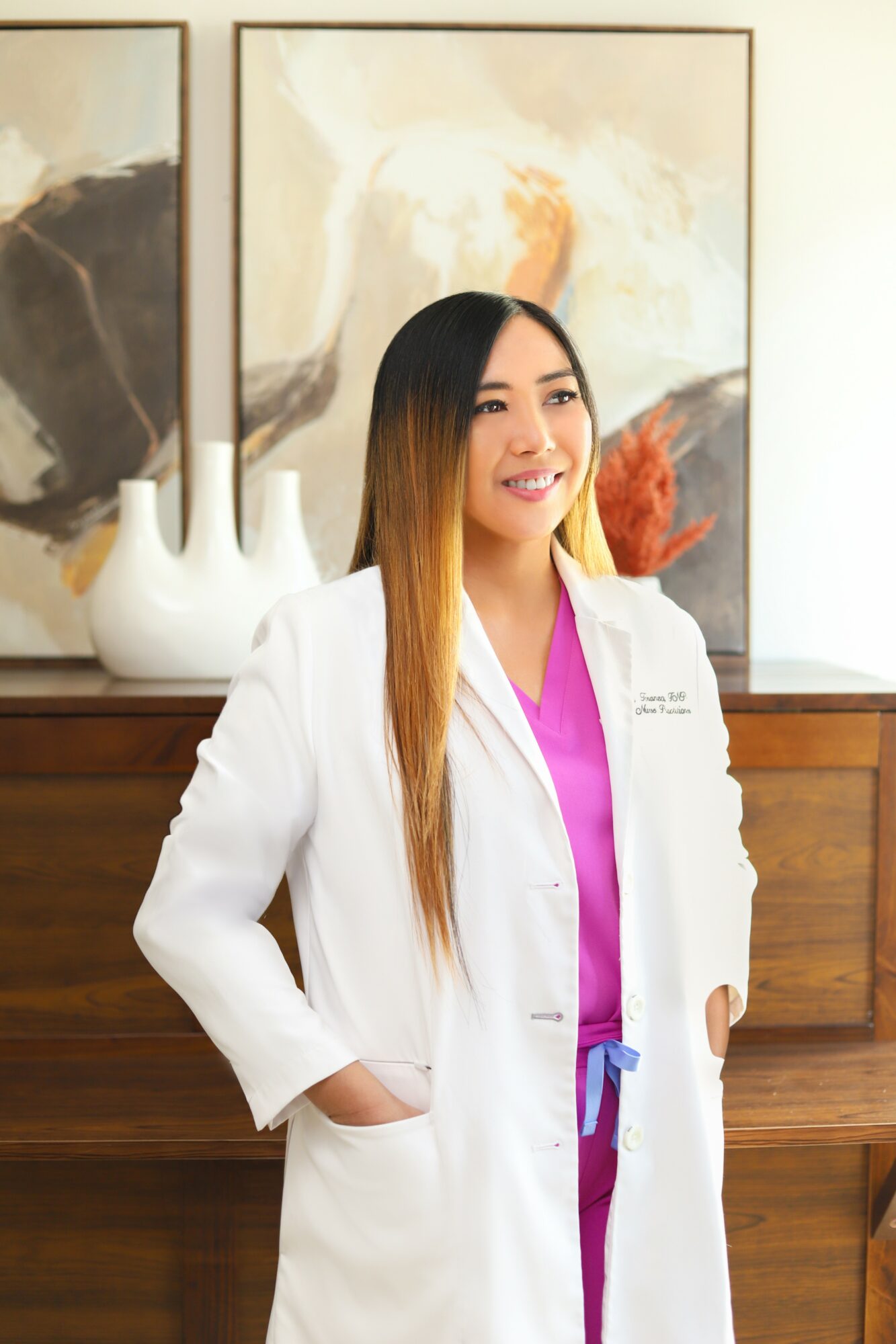 Woman in white coat and pink scrubs standing indoors with artwork and vases behind her.