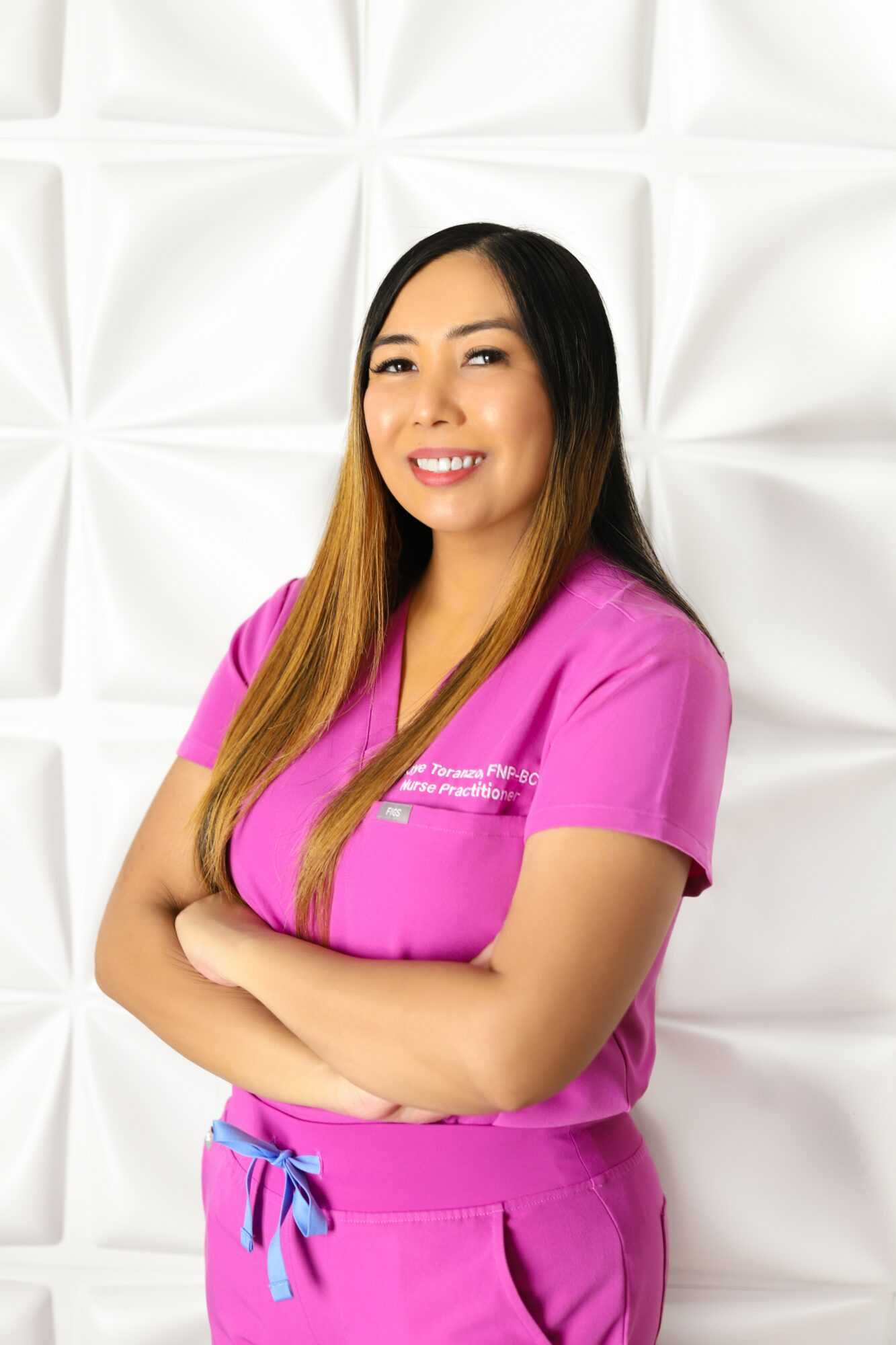 Woman in pink medical scrubs smiling with arms crossed, standing against a white textured background.