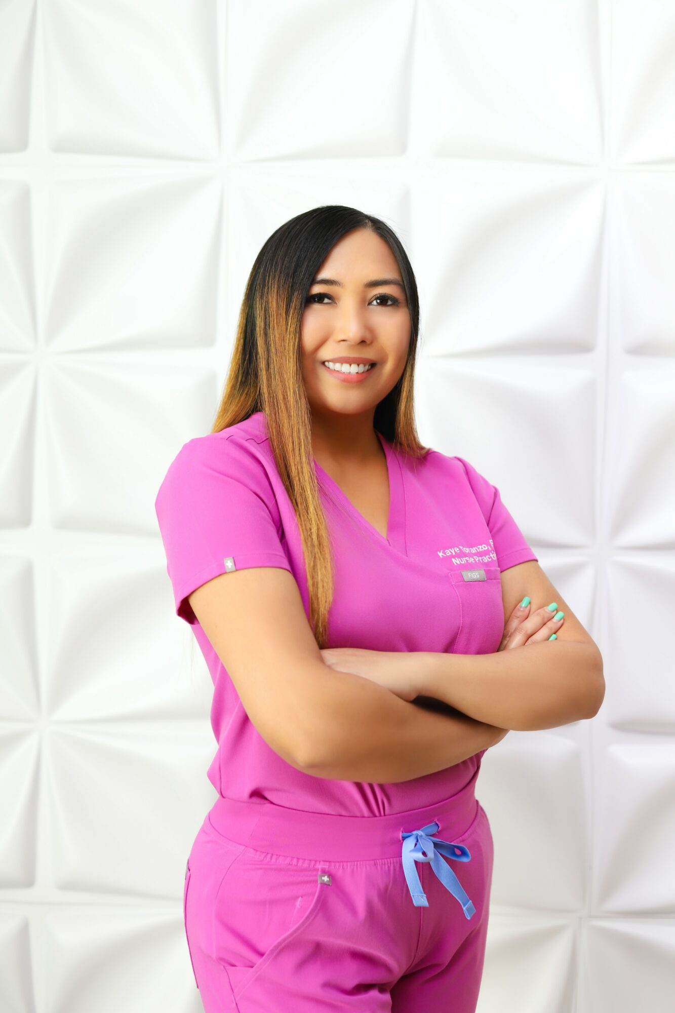 Young woman in pink medical scrubs smiling with arms crossed, standing against a white textured wall.
