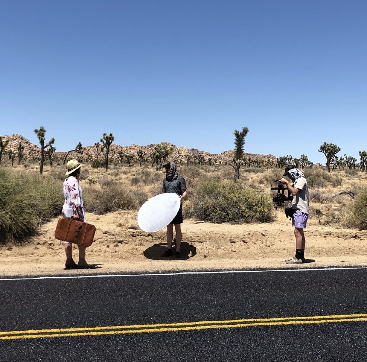 Three people on a desert roadside, one with a large umbrella, one with a camera, and one with a bag, desert landscape.