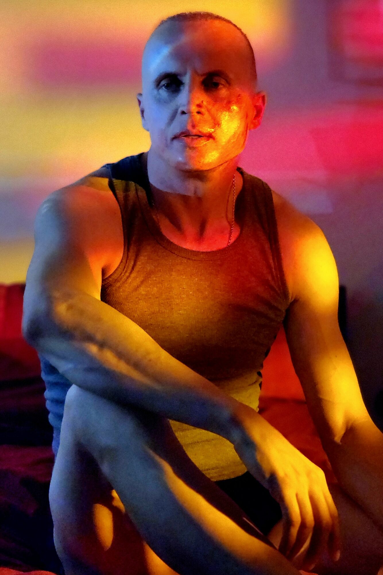 Person with shaved head and muscular arms, wearing a sleeveless top, in colorful lighting background.