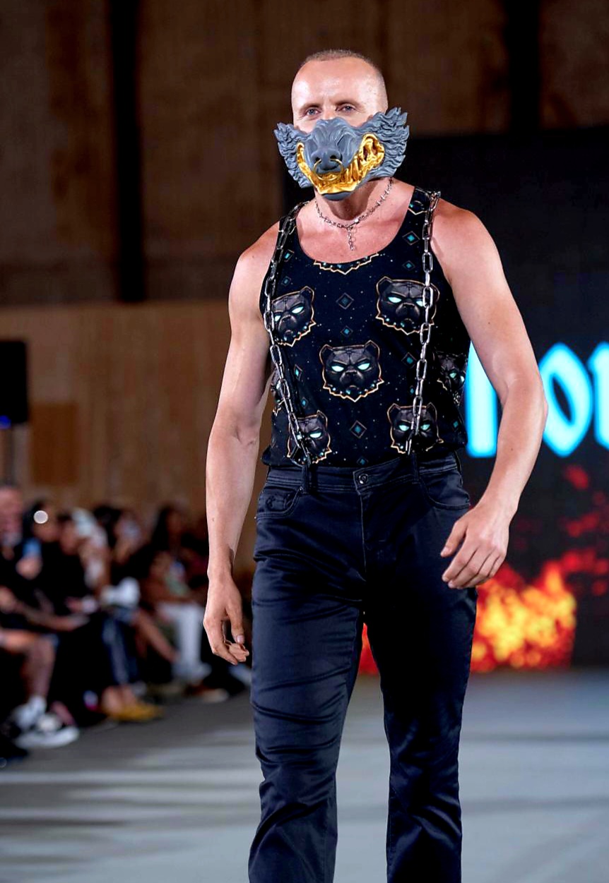 Person walking on runway wearing a mask with a blue and yellow design, black sleeveless top, and dark pants.